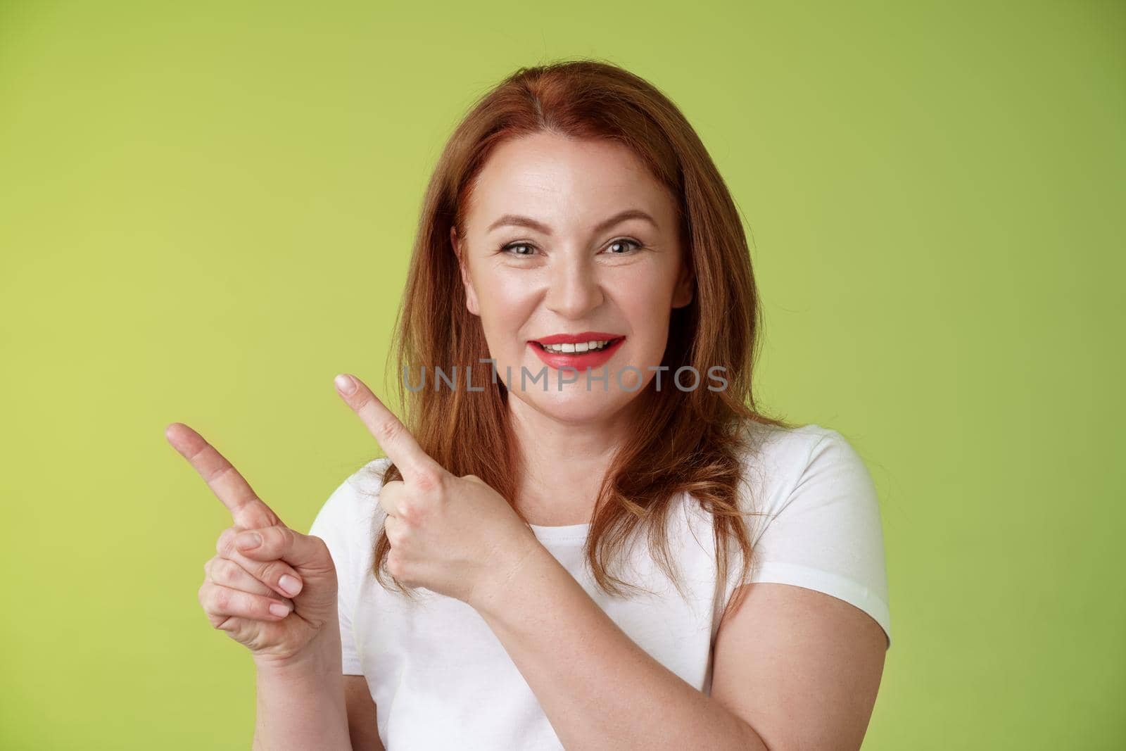 Close-up joyful motivated pleasant redhead middle-aged female. pointing upper left corner index fingers smiling delighted give advice check-out promo good advertisement blank space green background.