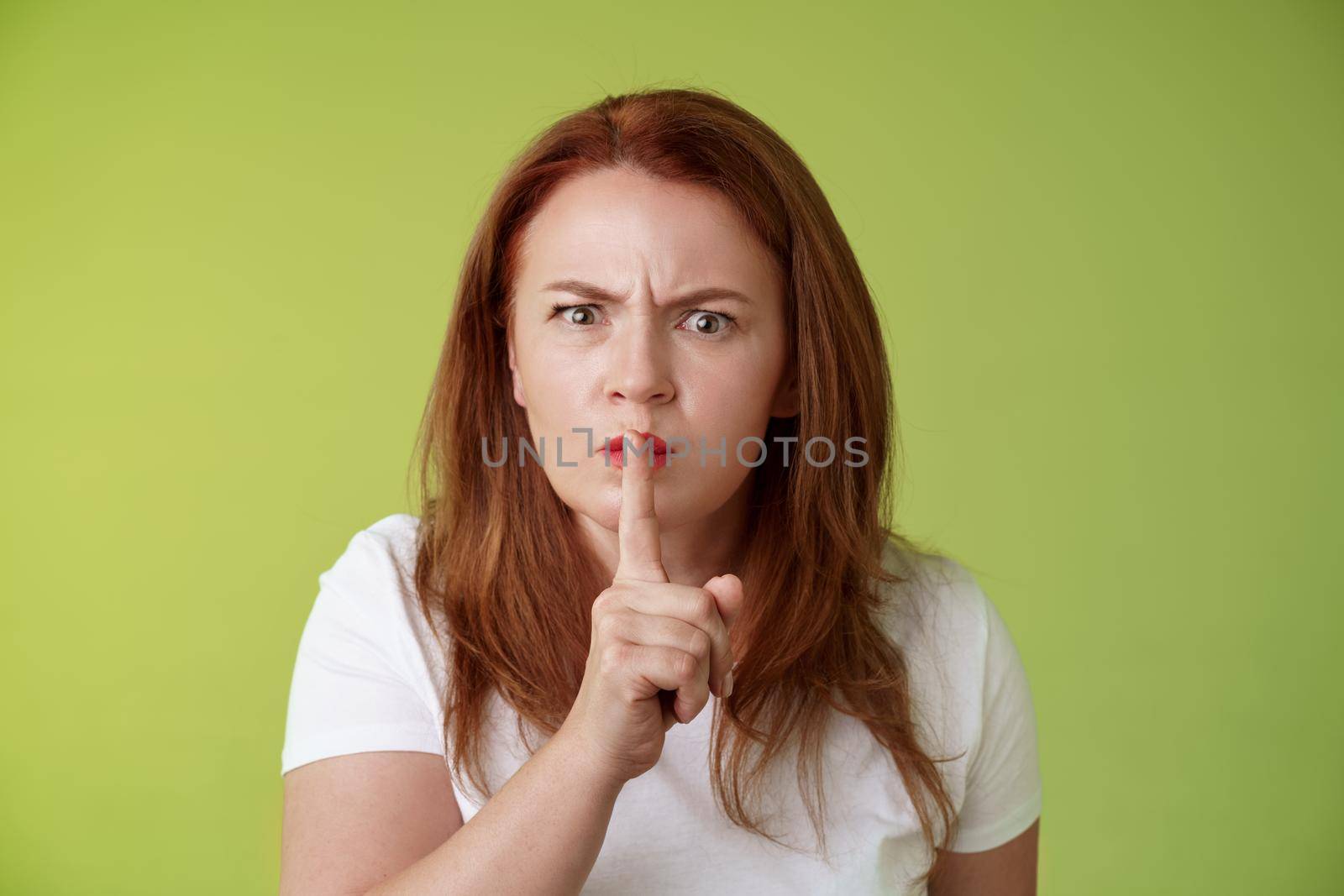 Not speak during exam. Strict serious-looking displeased middle-aged redhead woman frowning disappointed hushing say shush index finger pressed lips keep quiet gesture green background.