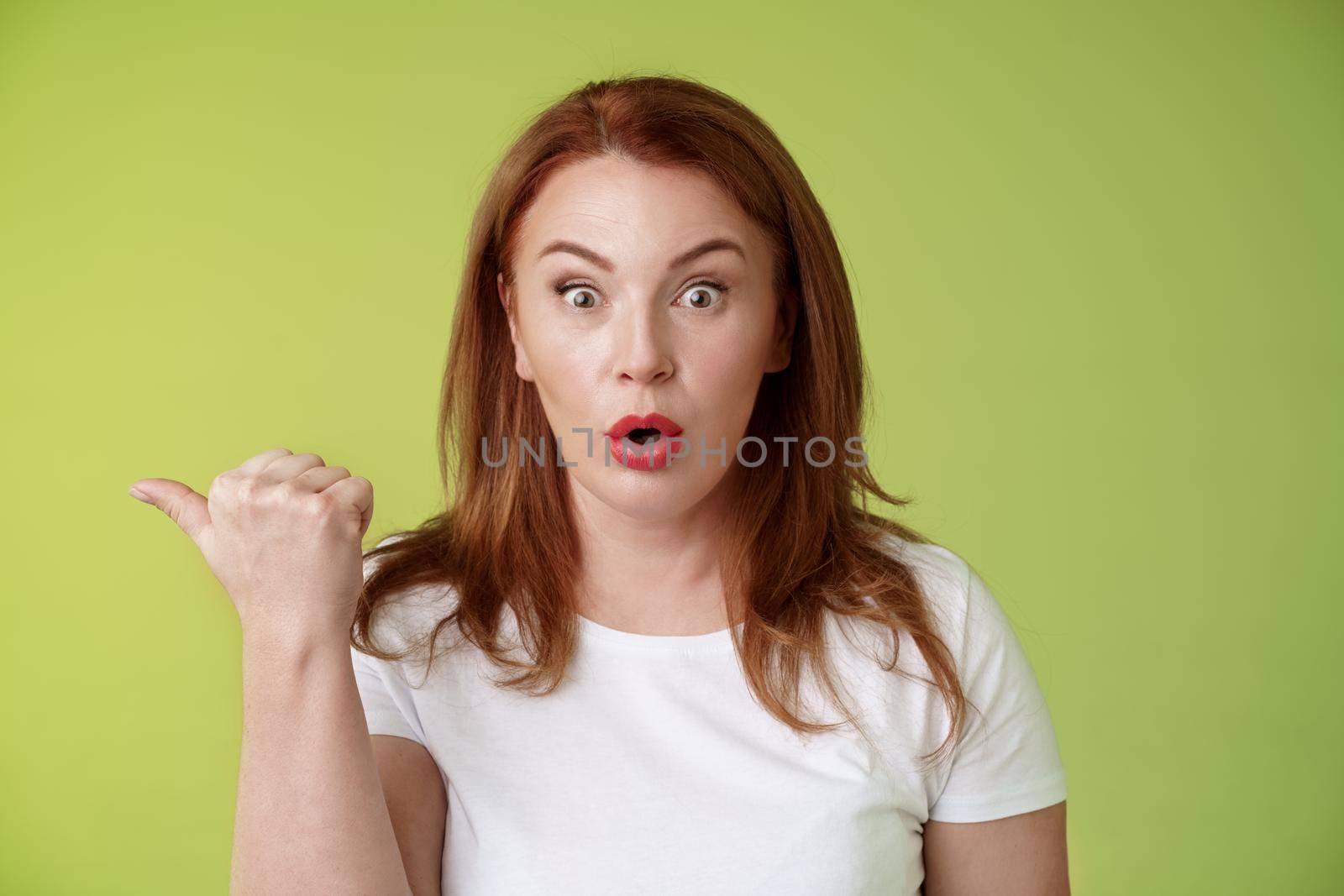 Close-up surprised amused redhead middle-aged housewife. folding lips say wow impressed stare camera questioned ambushed pointing left thumb astonished wondering if promo true green background.
