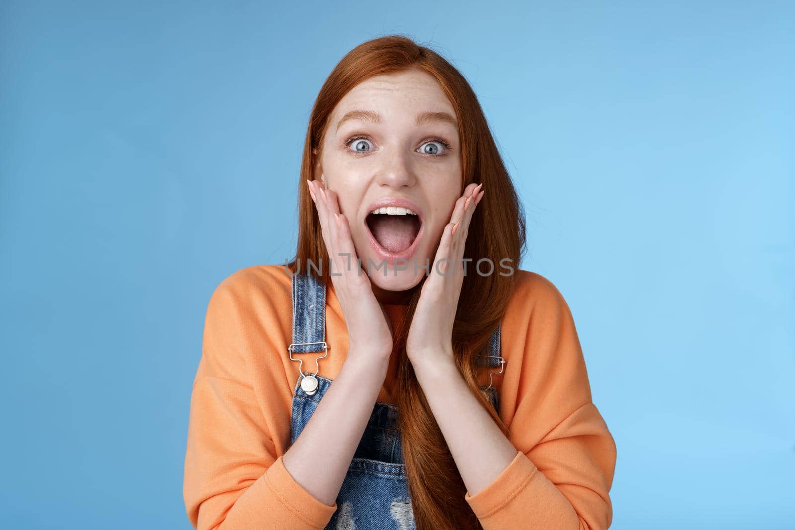 Excited thrilled young emotional enthusiasitc ginger girl teenage college student yelling amused smiling broadly receive positive good news look surprised camera touch face astonished blue background by Benzoix
