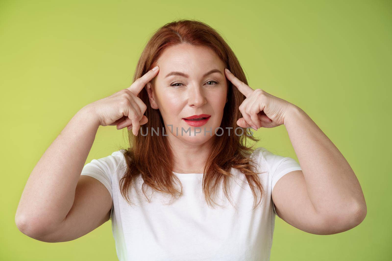 Smart choice. Intrigued thoughtful good-looking assertive redhead creative middle-aged woman touch temples squinting pondering info thinking use mental power read minds stand green background by Benzoix