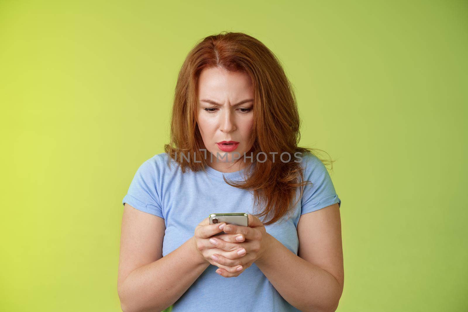Confused unsure redhead middle-aged woman learn how use social media trying understand emoji look intense focused smartphone display reading important news message stand green background by Benzoix