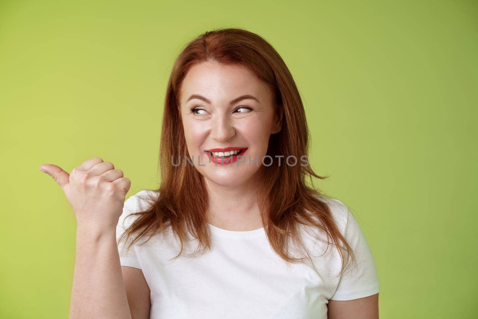 Intrigued charismatic redhead middle-aged woman smiling temtation interest pointing looking left curiously check-out cool promo share awesome place location stand green background.
