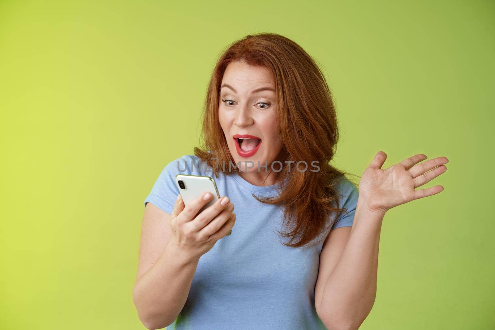 Surprised glad middle-aged pretty redhead woman look smartphone raise hand celebratory excitement open mouth. reacting impressed gladly read good excellent news look mobile screen green background.