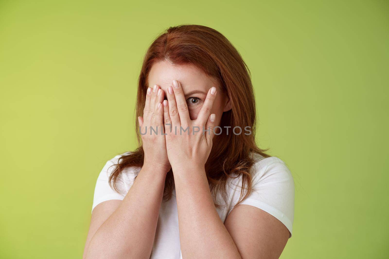 Not peeking. Playful charismatic middle-aged ginger redhead woman close face palms look through fingers one eye stare camera intrigued waiting surprise gift stand green background by Benzoix