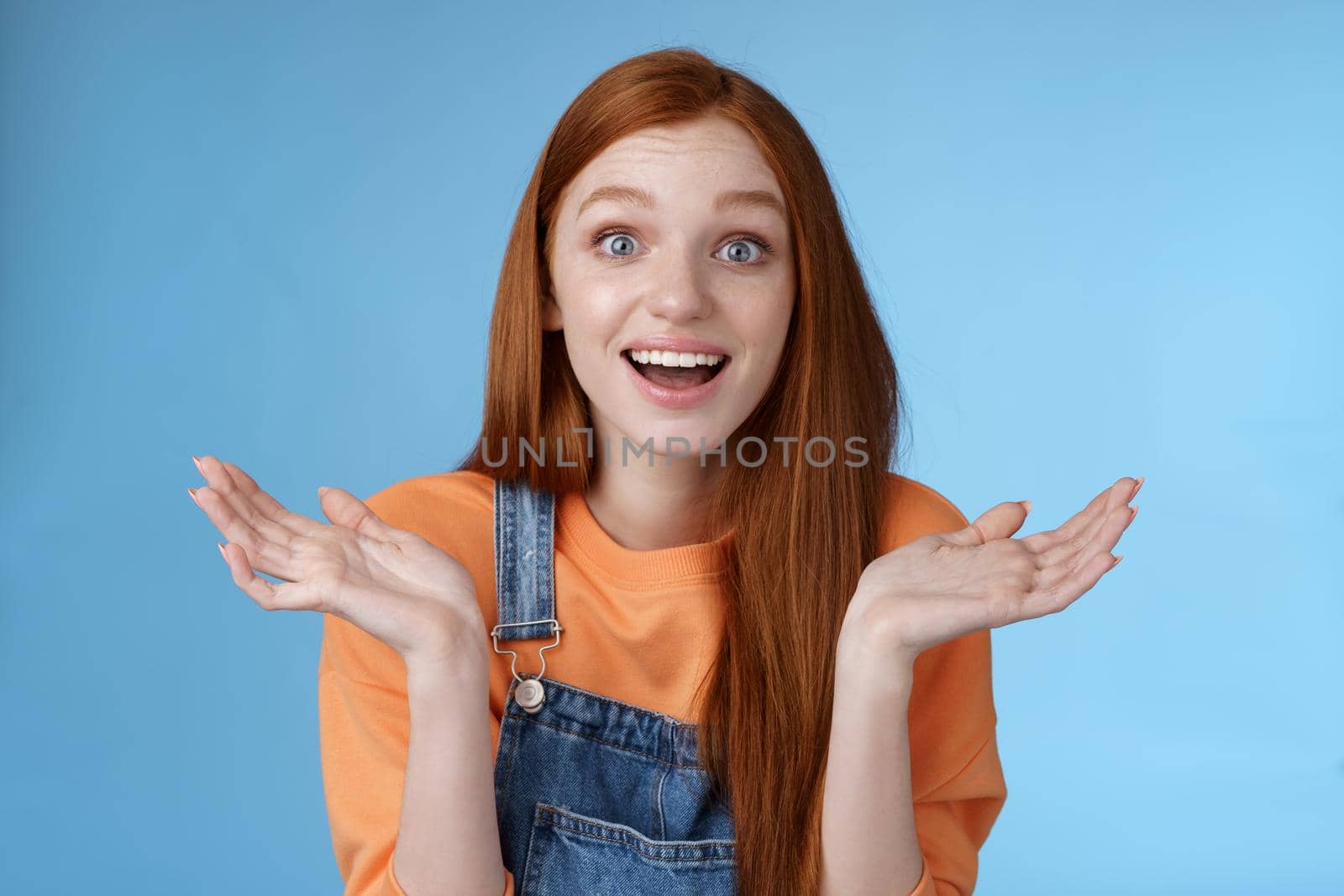 Amused surprised glad young redhead girl speechless happy see friend came back hometown wide eyes impressed grinning raise hands sideways full disbelief standing blue background joyful by Benzoix