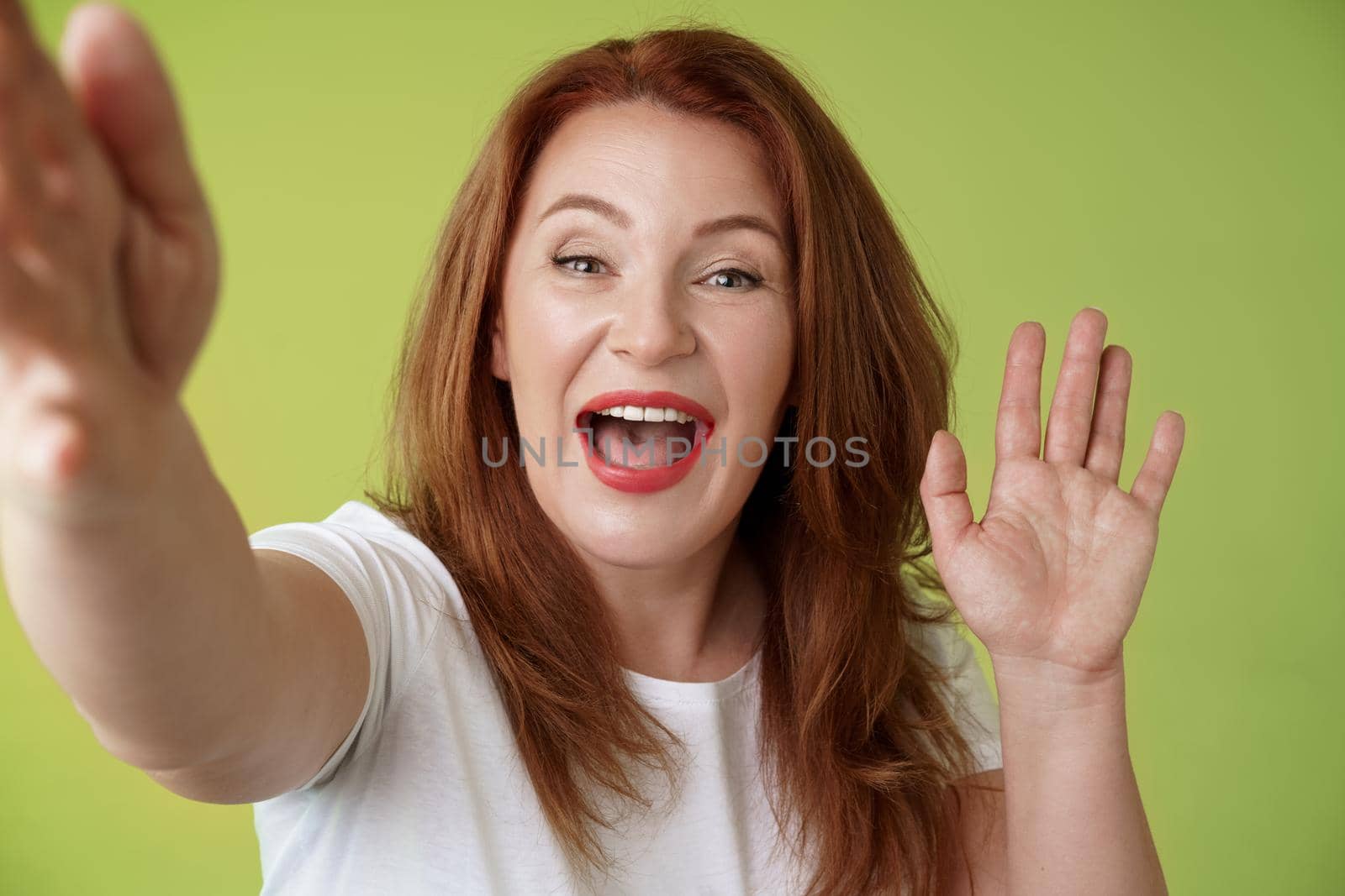 Friendly cheerful redhead middle-aged woman. extend arm hold camera taking selfie waving palm hi hello greeting smiling broadly welcome daughter talking video-call mobile internet green background.