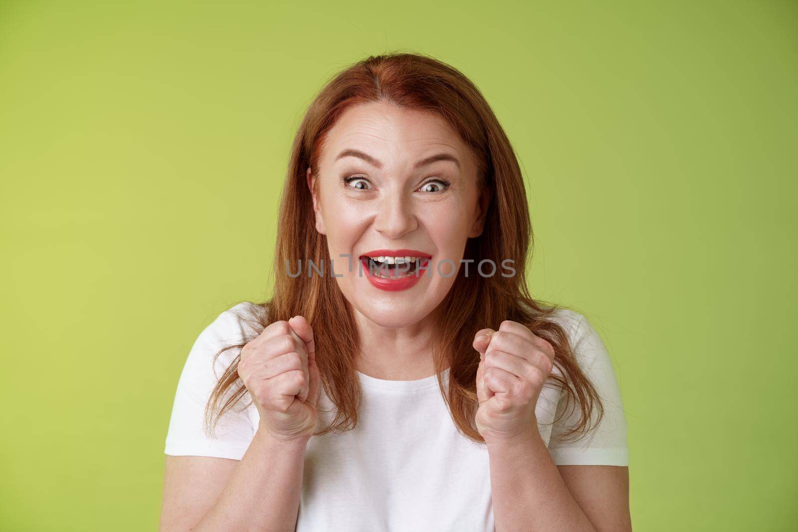 Close-up lucky enthusiastic cute redhead joyful middle-aged woman. pump fists vigorous excitement celebratory smiling broadly winning celebrating triumphing success good news standing green background.