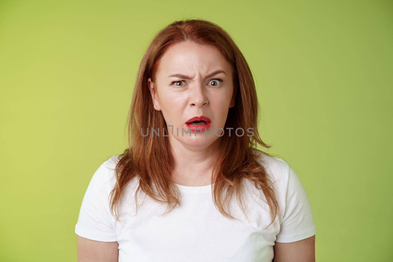 Confused shocked gasping middle-aged redhead woman cringe frustrated puzzled open mouth speechless freak out strange shocking scene stand green background perplexed disappointed by Benzoix