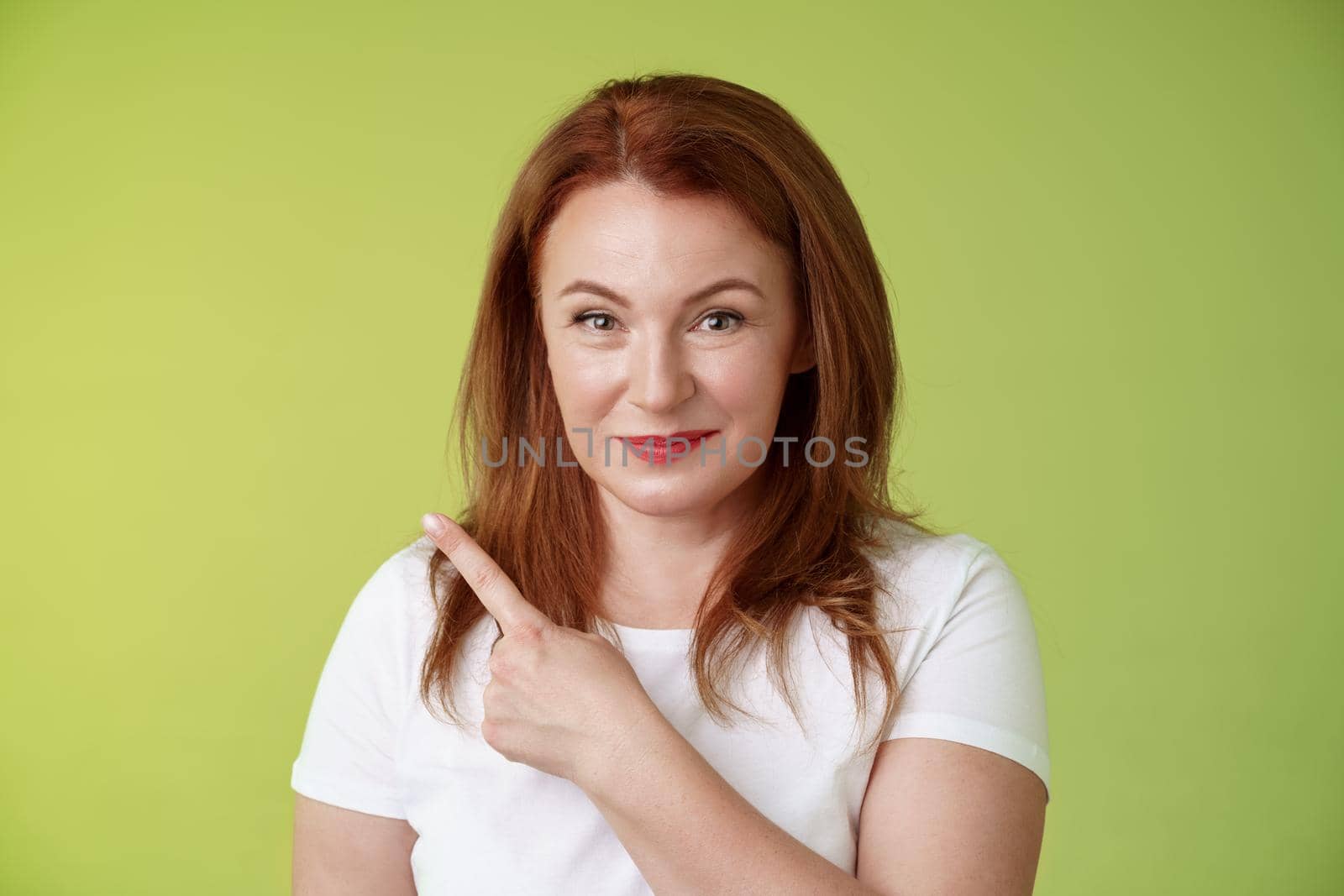 Kind cheerful good-looking middle-aged woman 50s redhead white t-shirt smiling modest assured give advice pointing upper left corner indicating great promotion advertising product green background by Benzoix