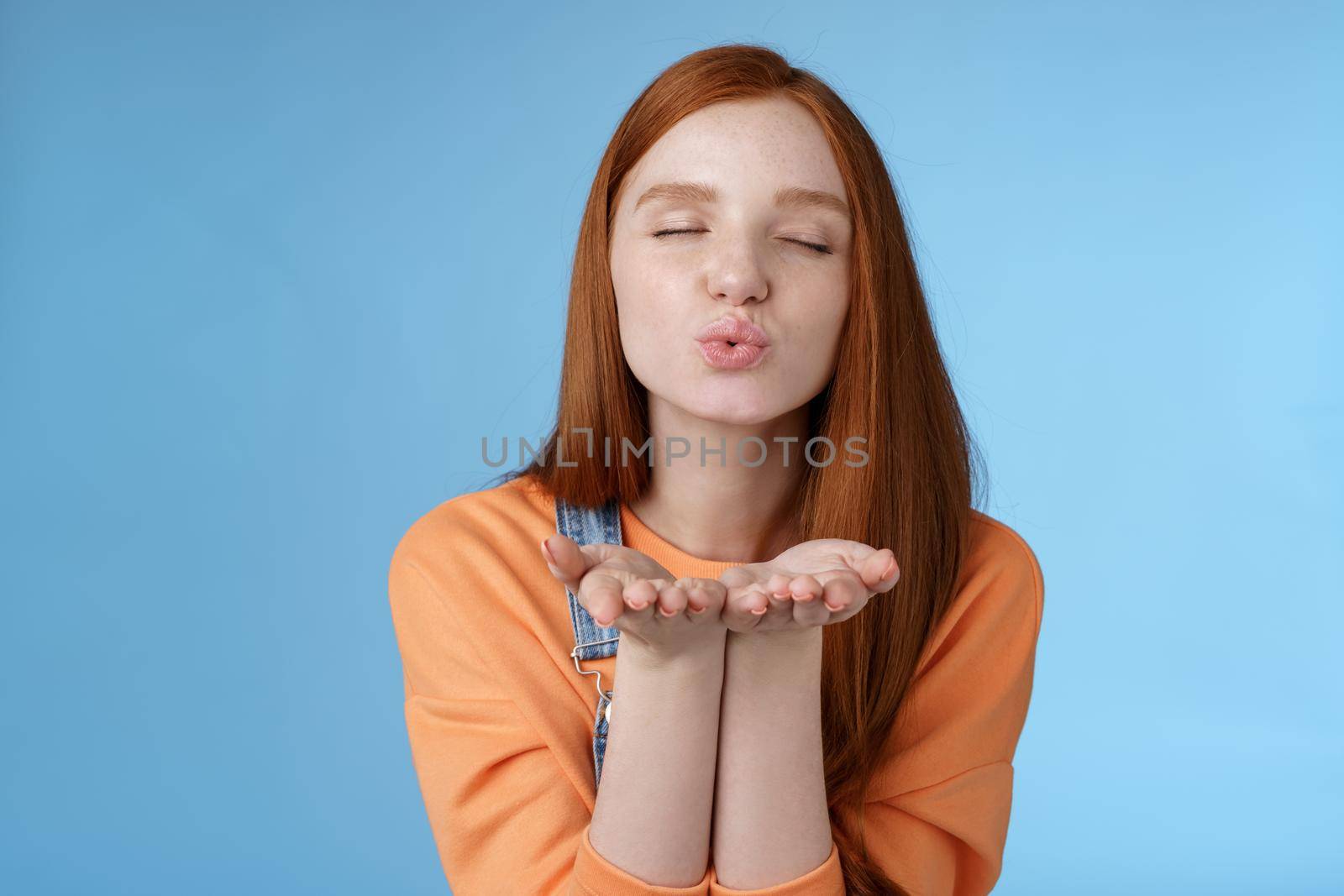 Passionate dreamy pretty redhead girl sending air kisses camera close eyes folding lips hold hands near mouth give muah internet followers recording vlog standing romantic tender blue background by Benzoix