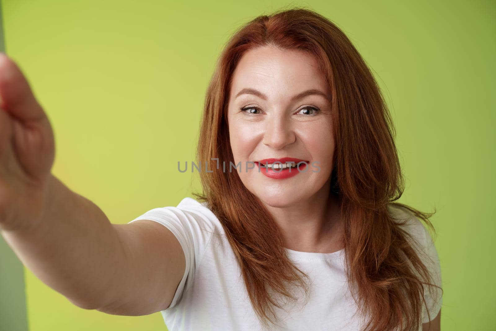 Close-up joyful enthusiastic redhead alluring middle-aged woman extand arm towards camera taking selfie smartphone smiling broadly posing photograph taking picture device green background.