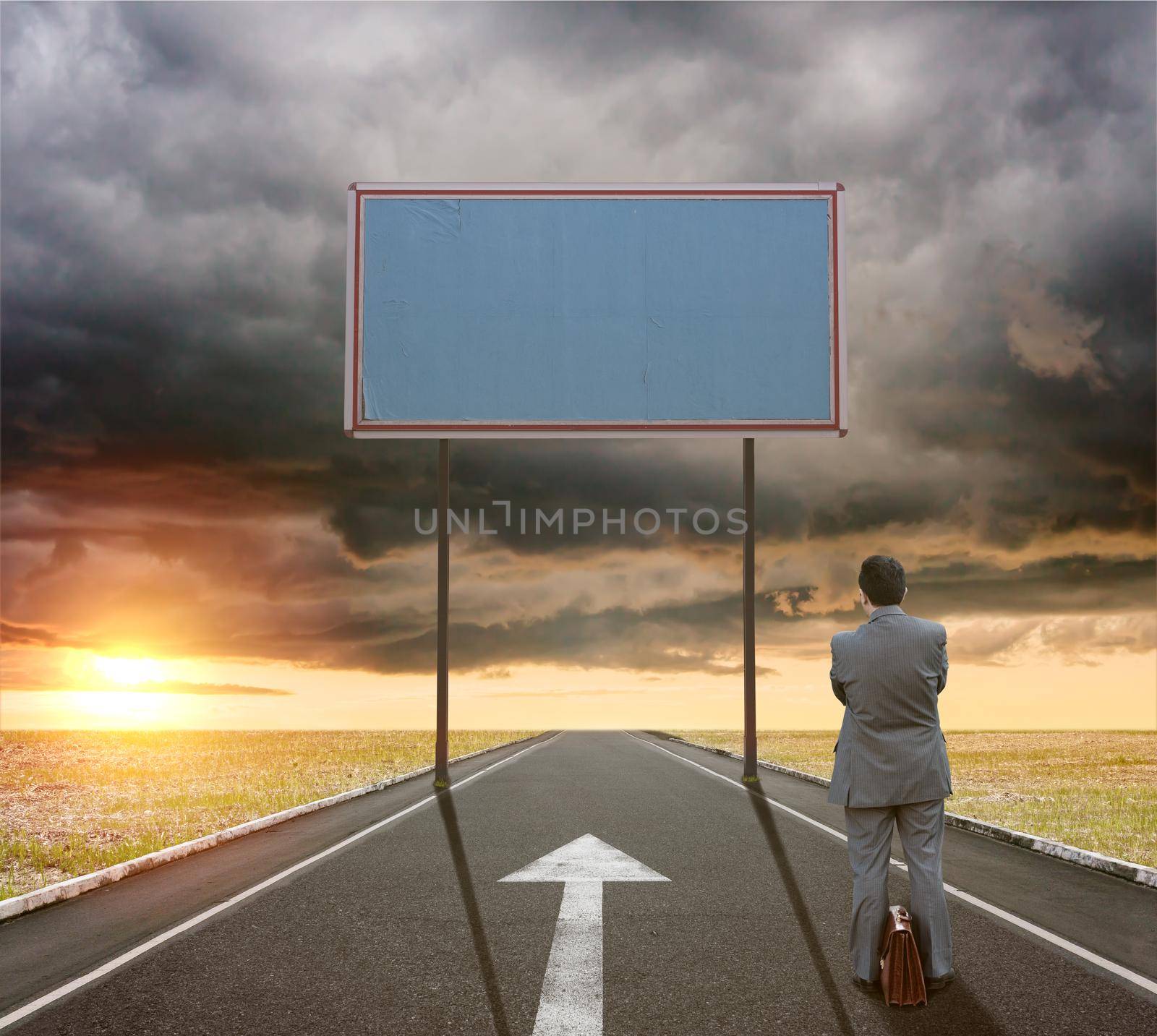 A businessman on the road next to a white arrow sign painted on the asphalt looks at a large empty billboard by bepsimage