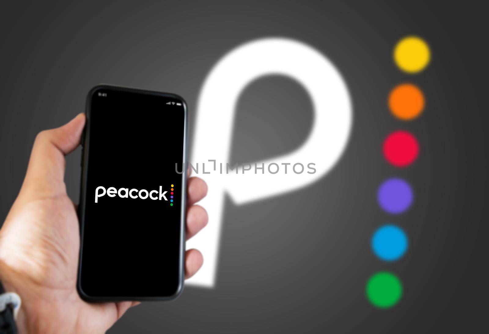 New York, USA, September 2021: hand holding a phone with the Peacock Tv mobile app on the screen and the Peacock Tv logo blurred on a grey background. Peacock is an American video streaming service