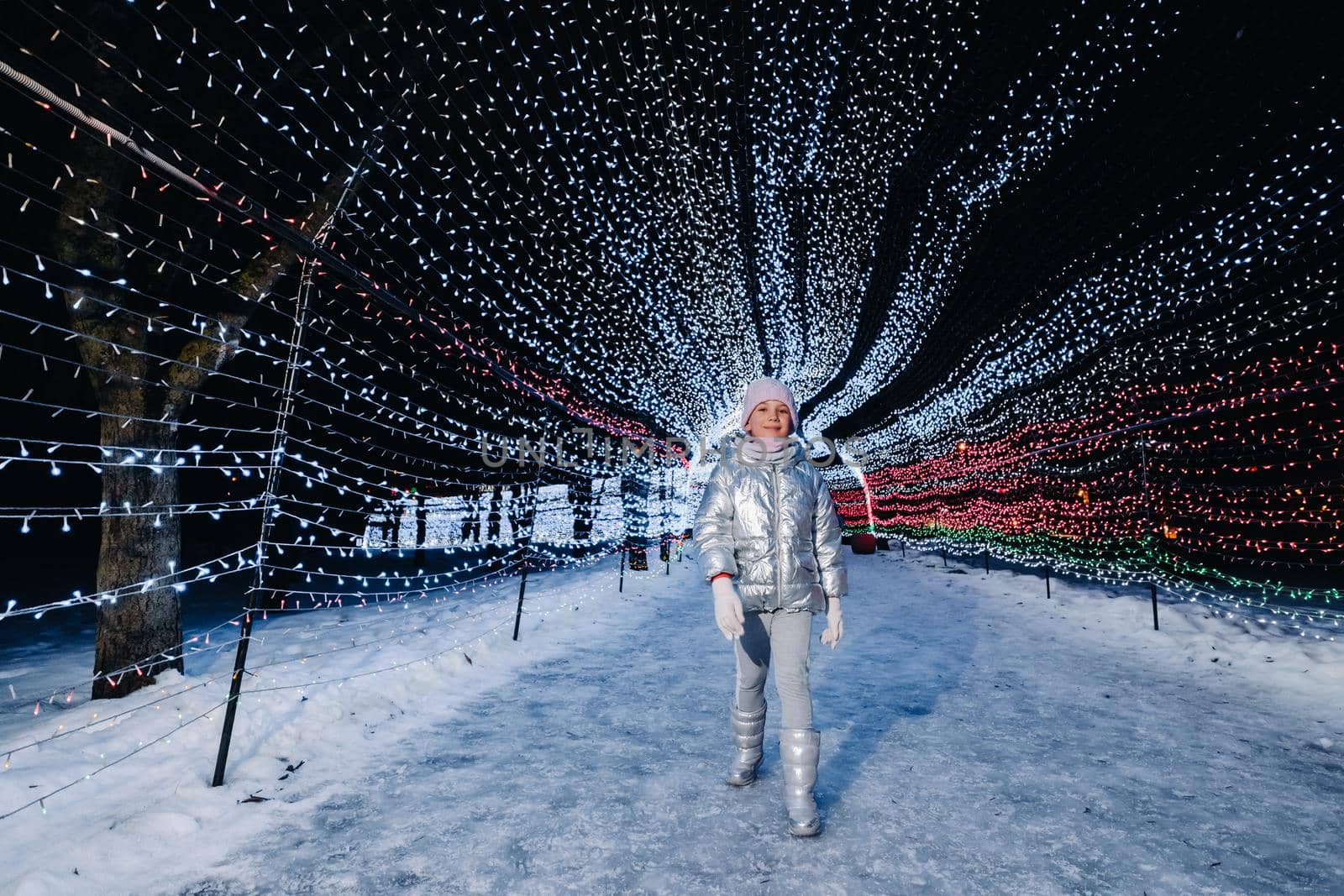 a little girl in a silver jacket in winter with evening burning lights on a Christmas decorated street.