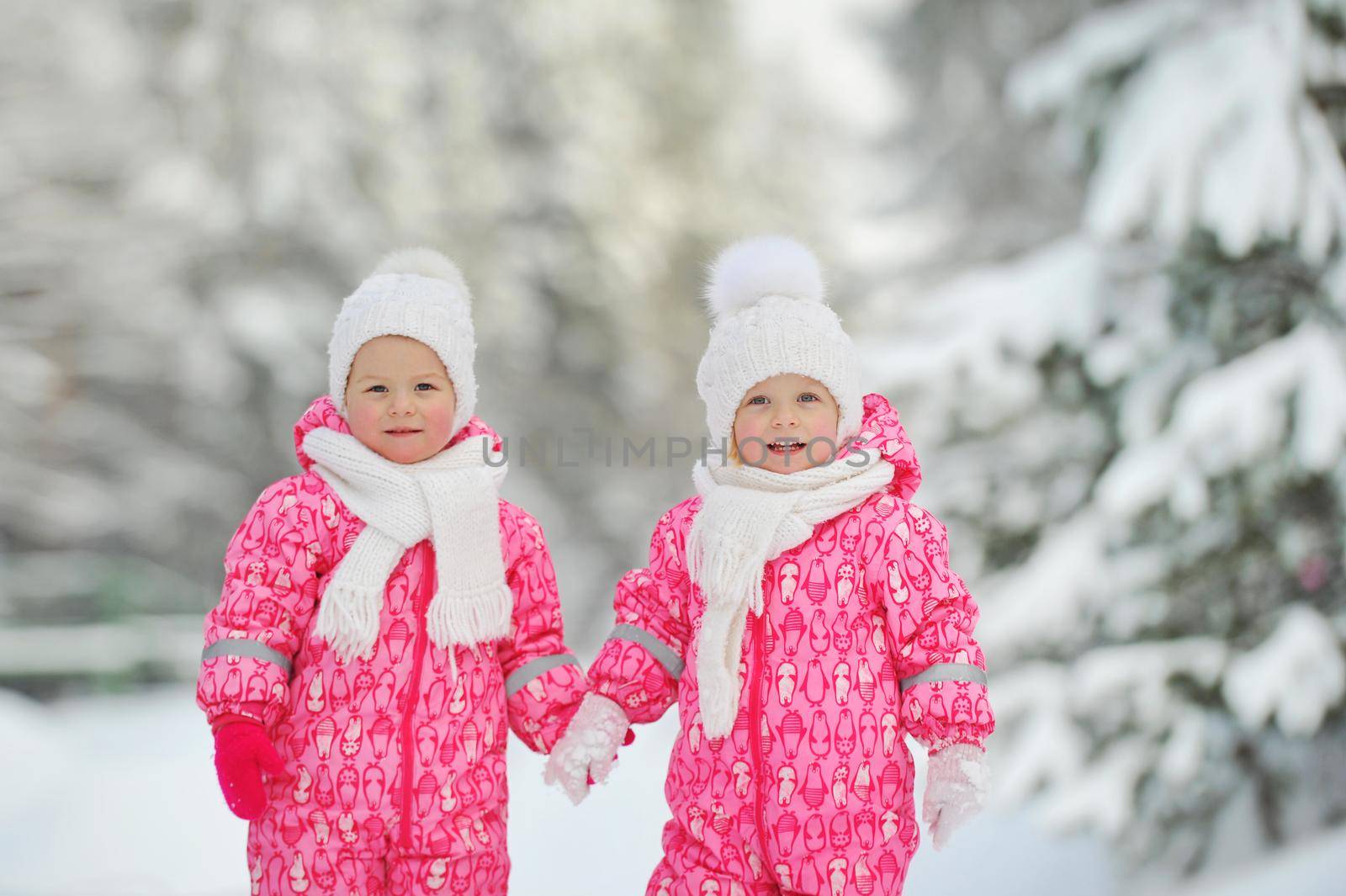 Two little twin girls in red suits stand in a snowy winter forest.