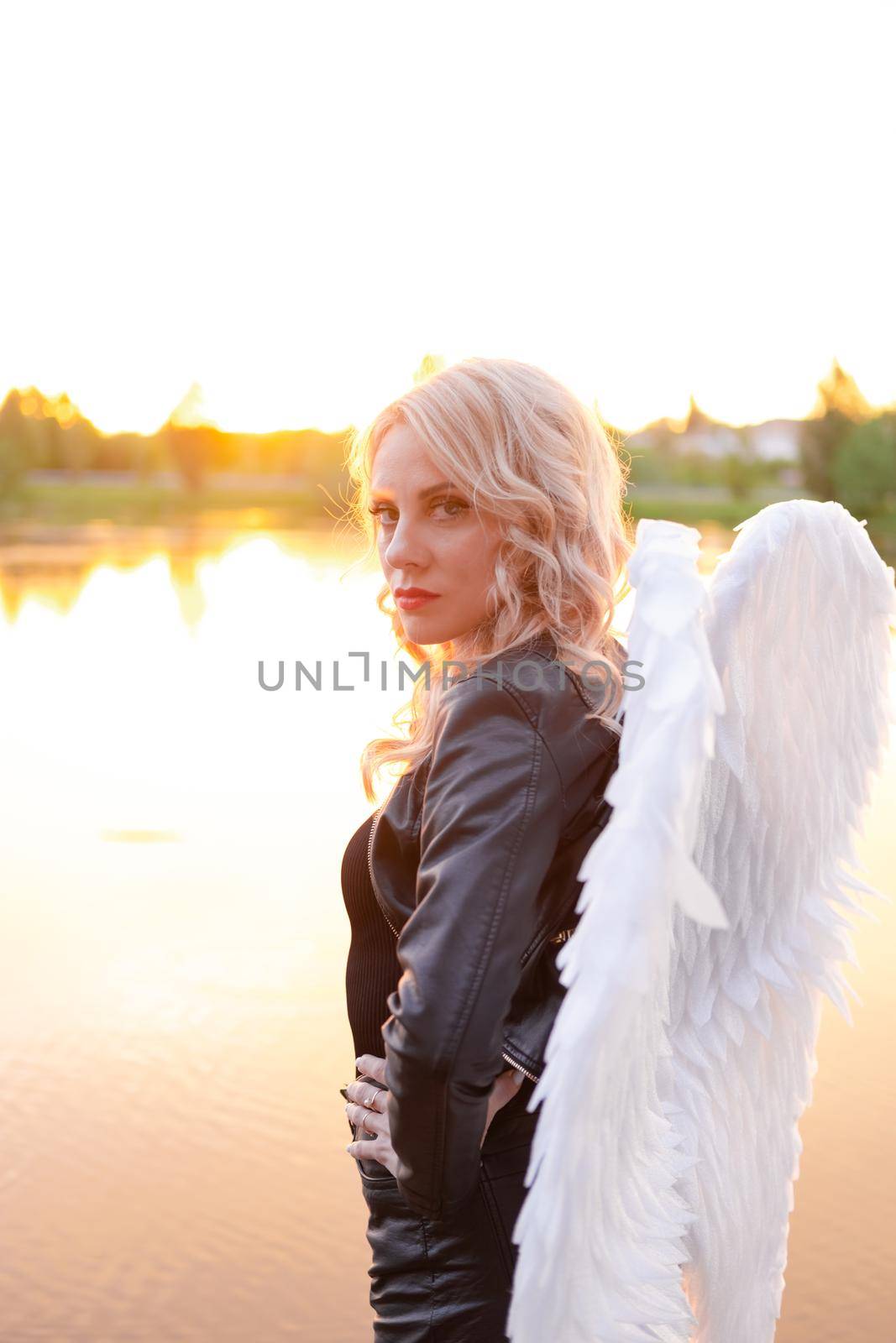sexy blonde woman in black leather jacket and shorts with white angel wings. demon or angel in hell or heaven. sunset near lake.