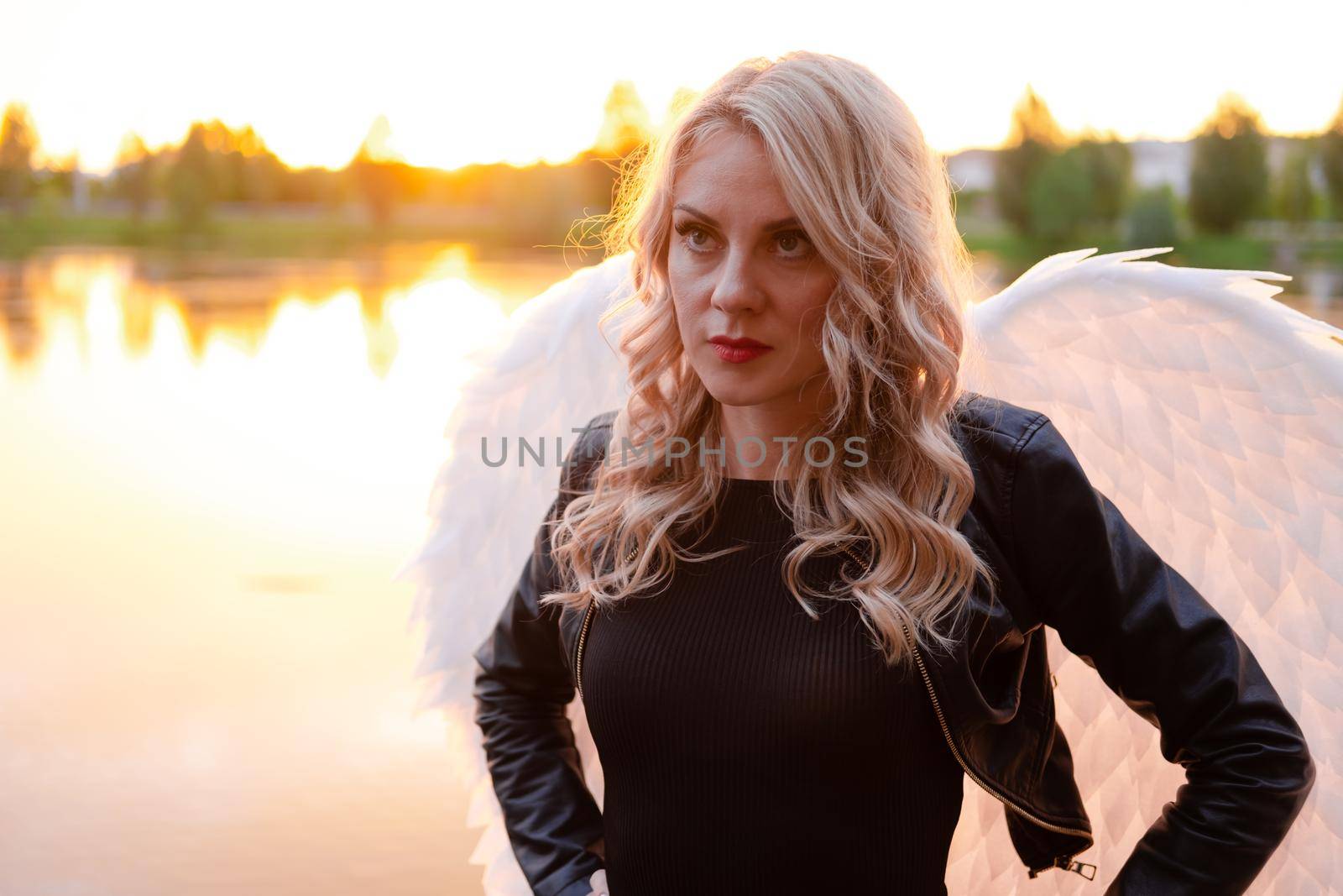 sexy blonde woman in black leather jacket and shorts with white angel wings. demon or angel in hell or heaven. sunset near lake.