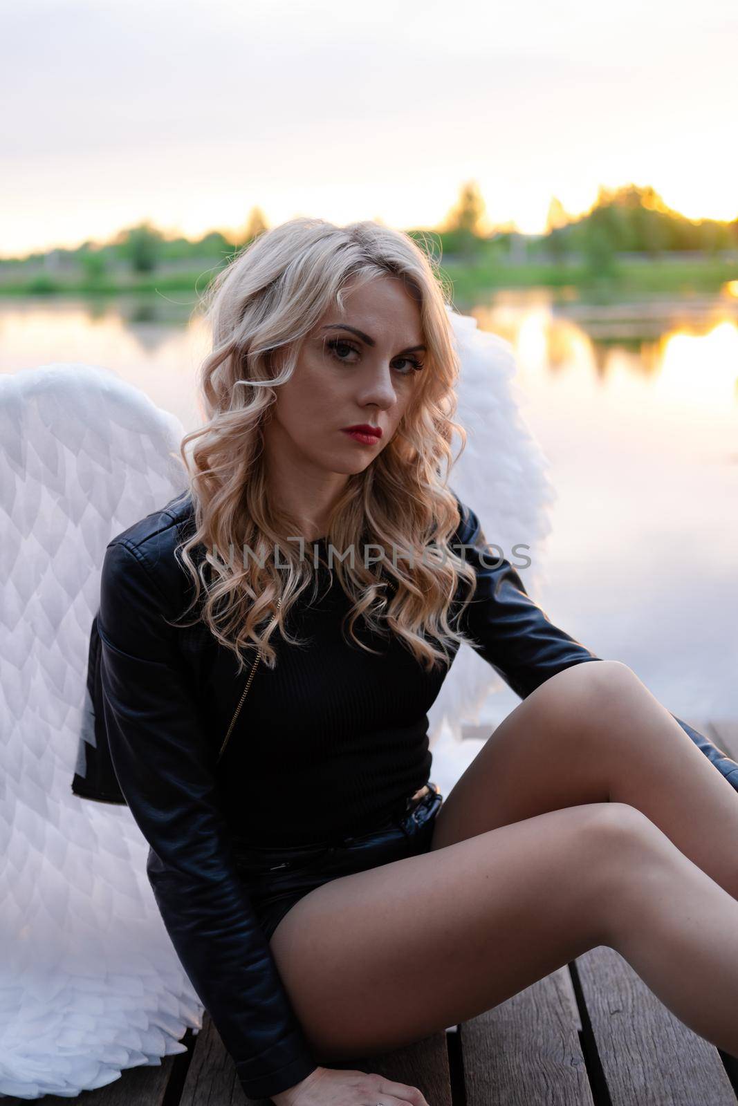 sexy blonde woman in black leather jacket and shorts with white angel wings. demon or angel in hell or heaven. sunset near lake by oliavesna