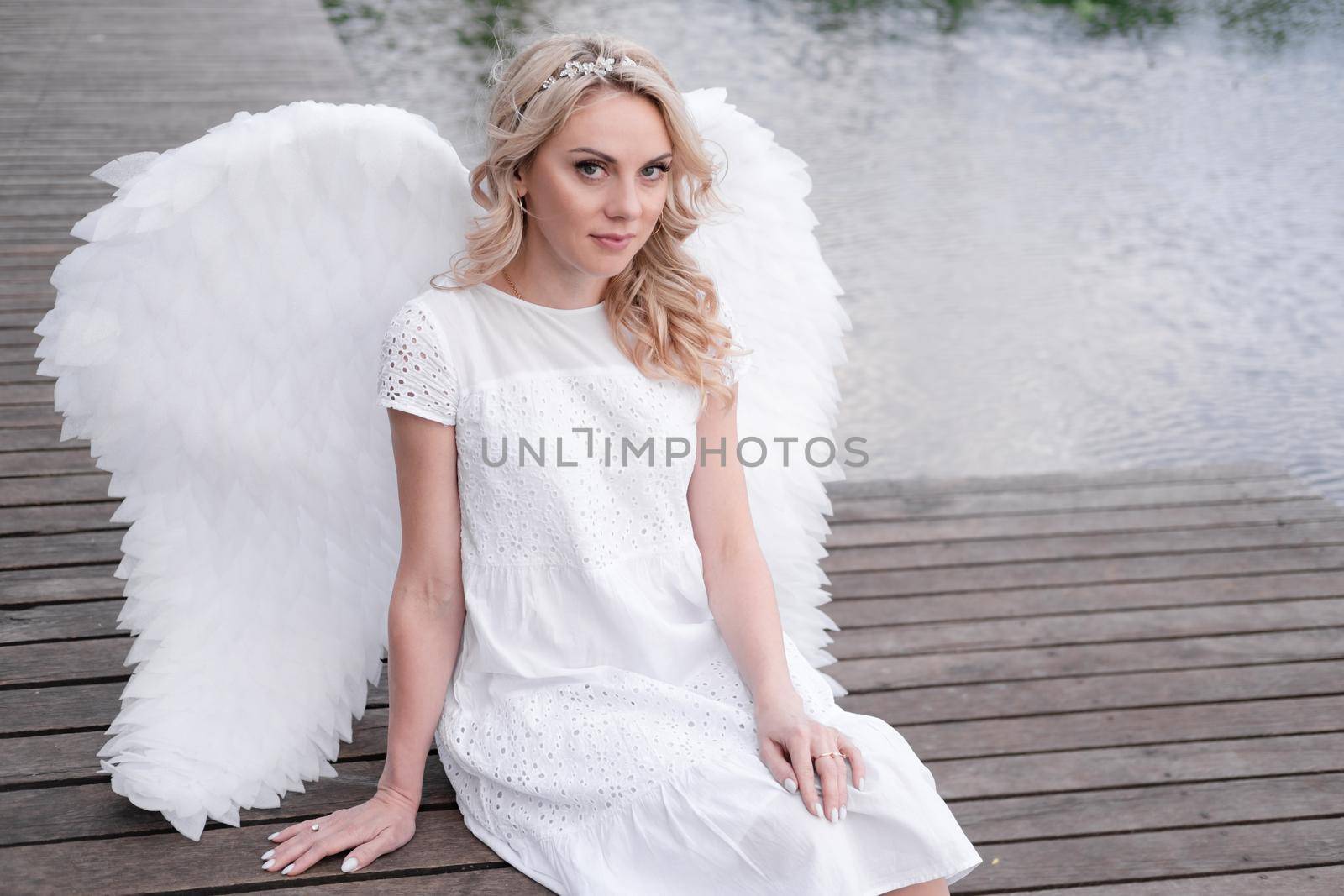 charming blonde with white angel wings standing by the water. beautiful woman in angel costume. goddess.