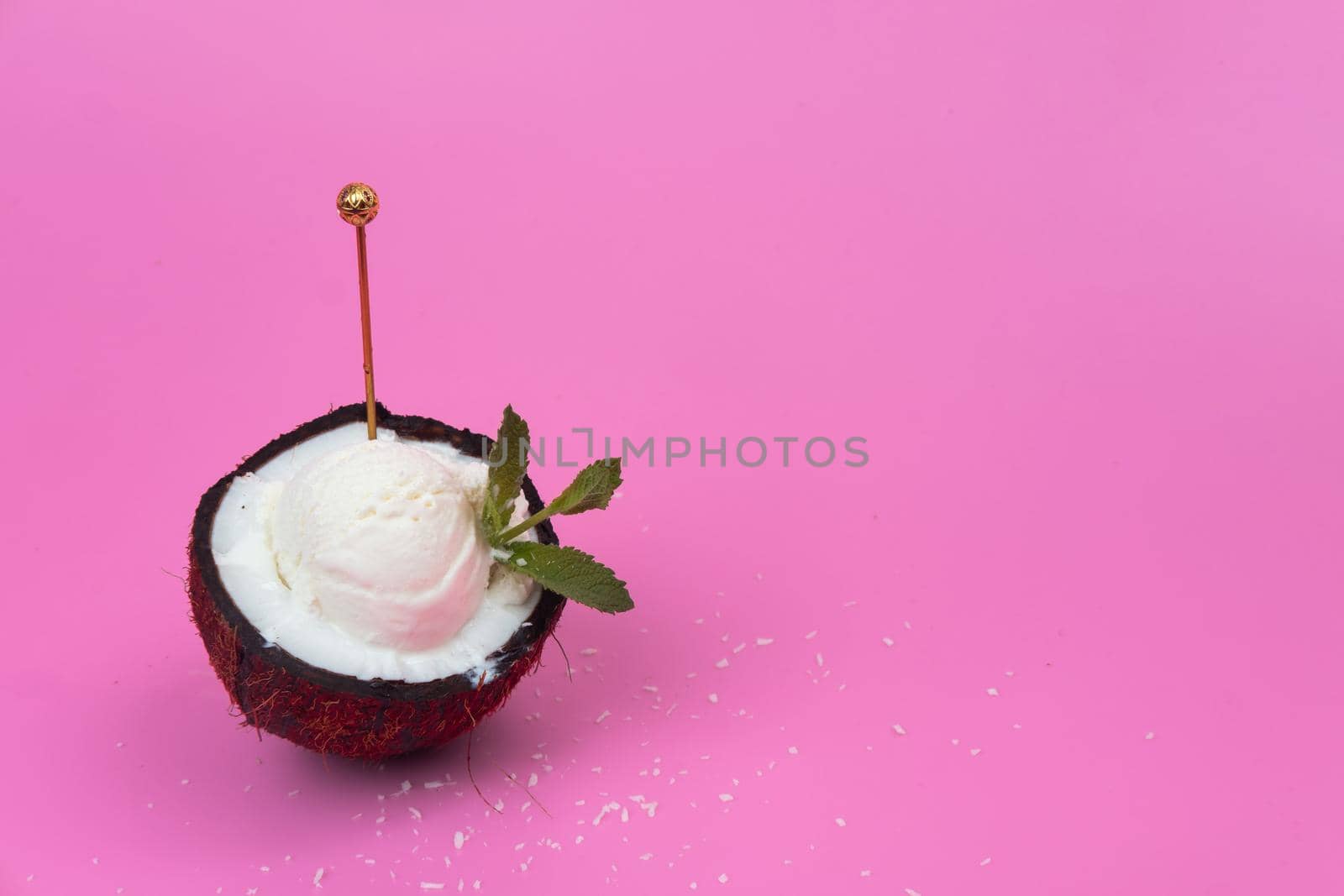 Vanilla ice cream balls in a fresh coconut half decorated with mint leaves with a spoon on a pink background.