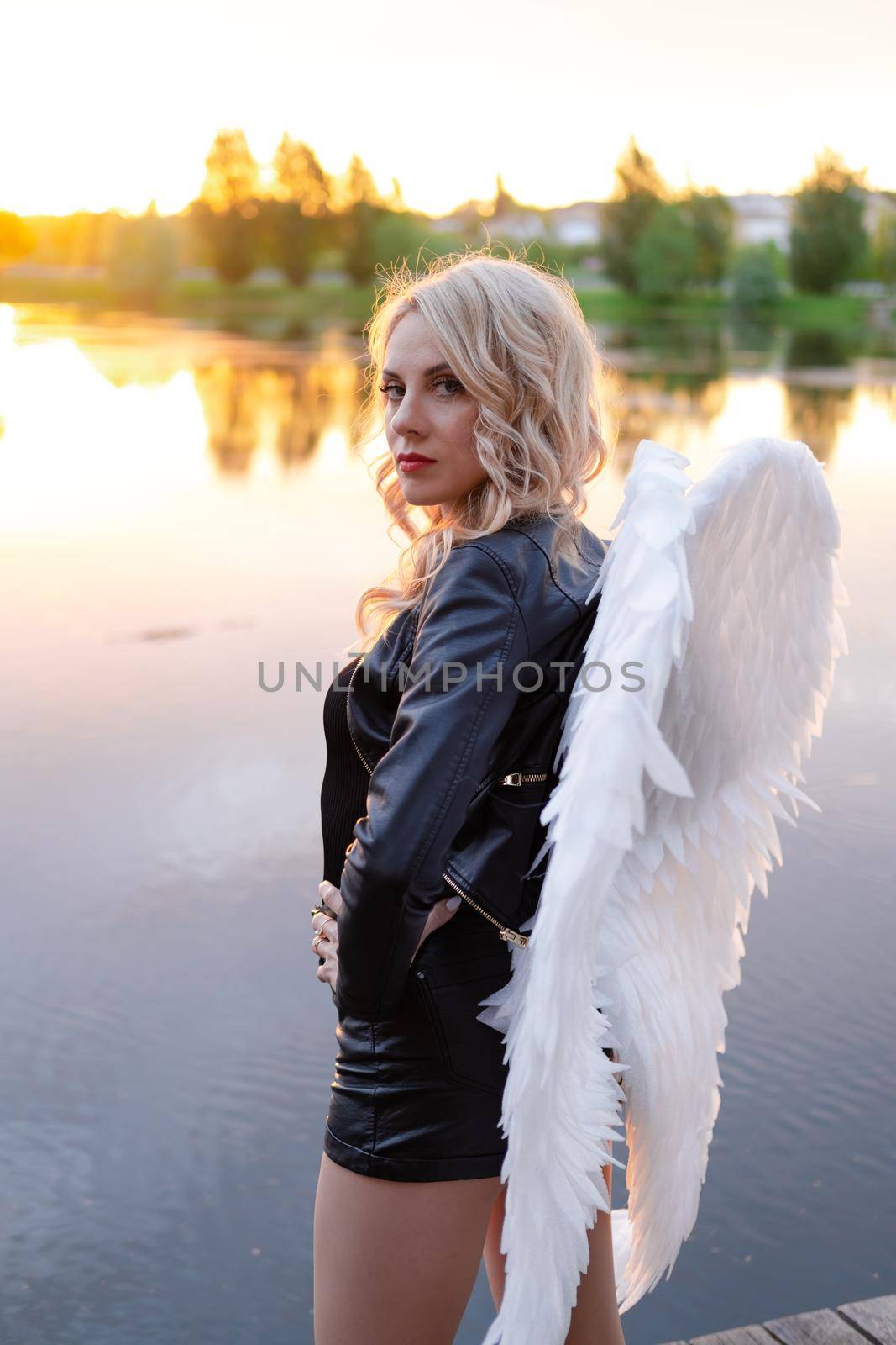 sexy blonde woman in black leather jacket and shorts with white angel wings. demon or angel in hell or heaven. sunset near lake by oliavesna