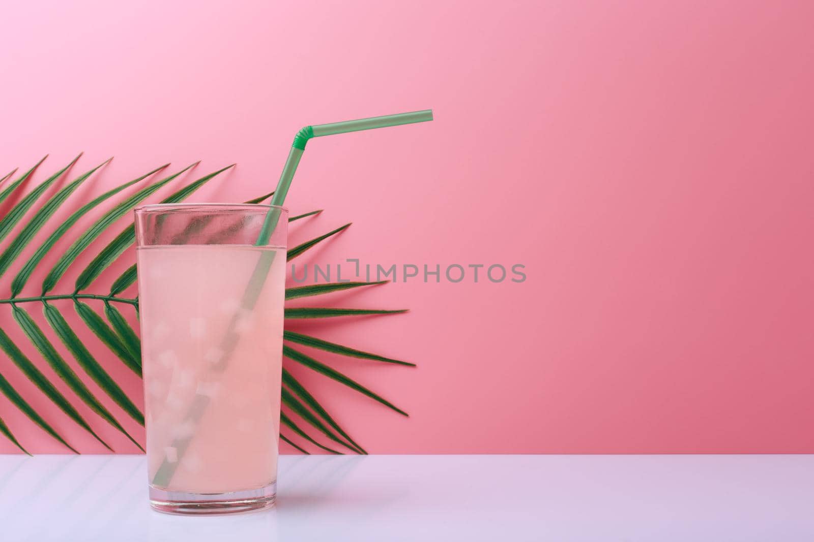 Creative summer composition with light pink fruit drink or lemonade with pieces of fruit and green straw on white table against pink background with palm leaf and copy space. Concept of refreshing cold drinks for summer or detox drinks