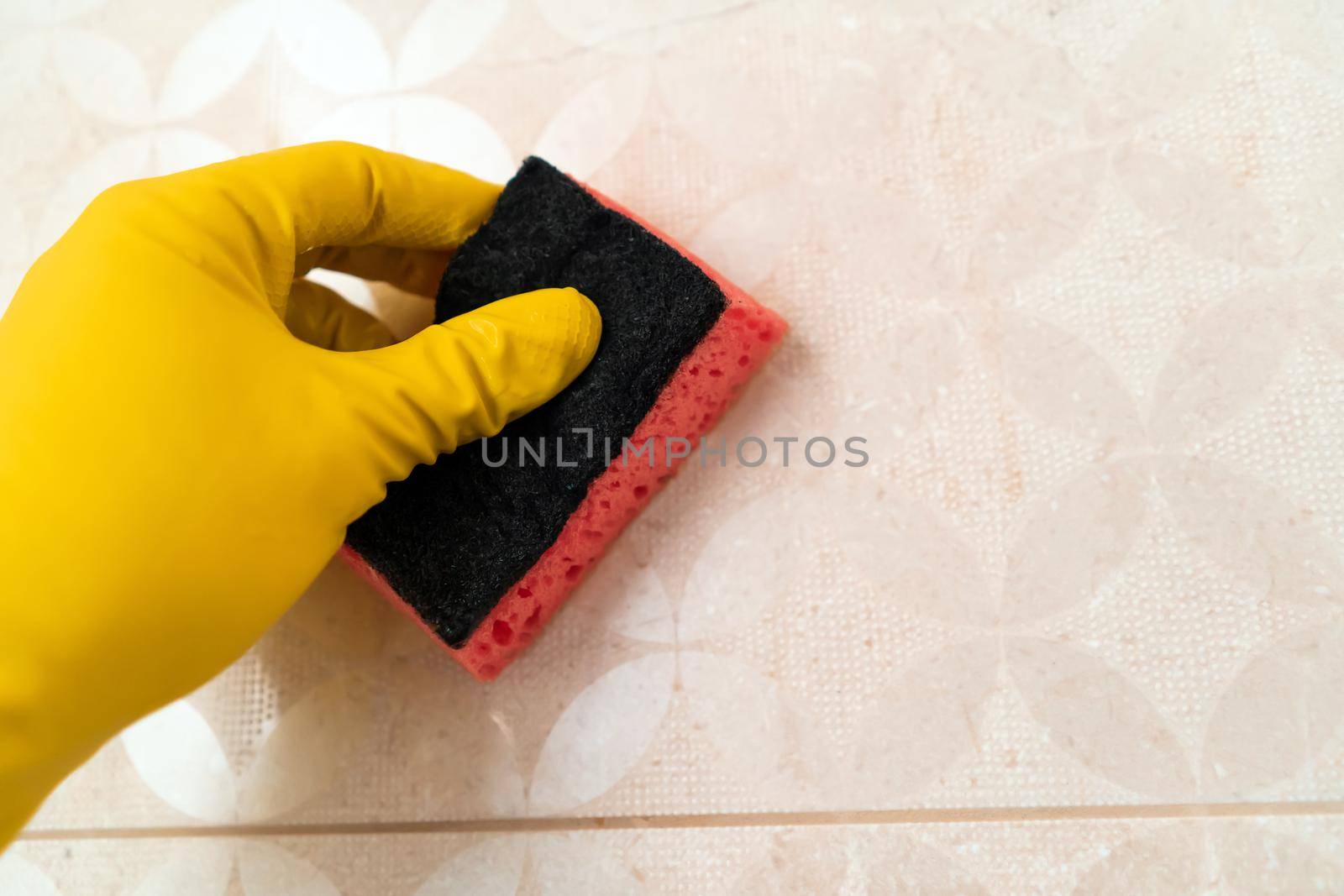 A hand in a yellow glove holds a sponge and wipes the tiles on the wall in the bathroom, a man does cleaning with detergent at home.