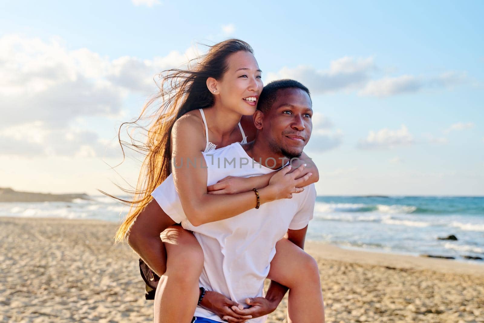 Young beautiful having fun couple on the sea background. Multiracial couple, asian woman on back of an african american man, vacation together, relationship, dating, happiness, happy lovers moments