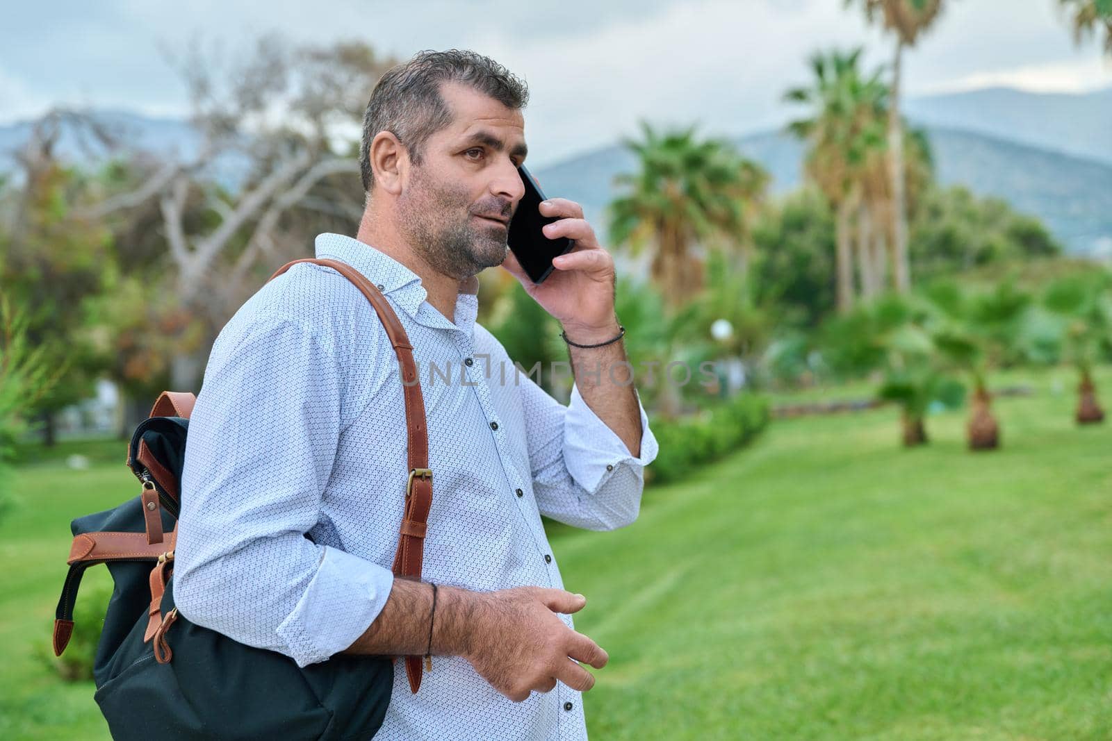 Mature business man talking on the phone outdoors. Serious confident male with backpack, in tropical park. Business trip, freelance, communication, technology, middle aged people concept, copy space