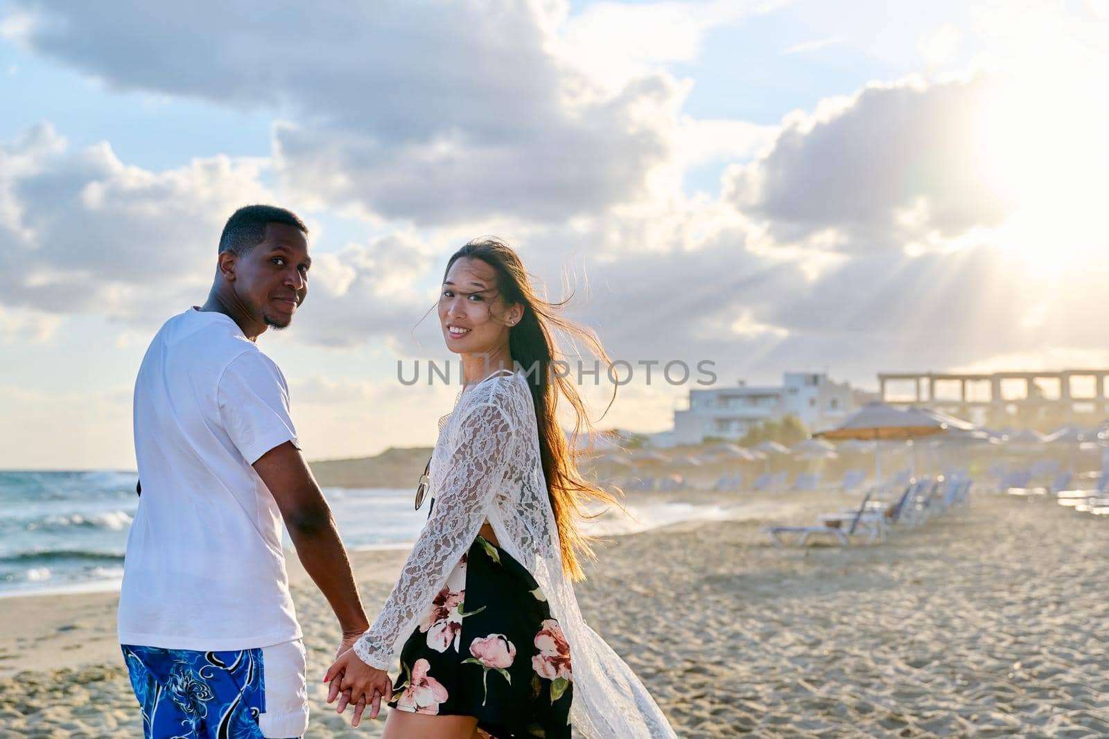 Portrait of beautiful young multiethnic couple holding hands on seashore. Happy romantic loving african man and asian woman looking in camera. People, love, relationship,multicultural family concept