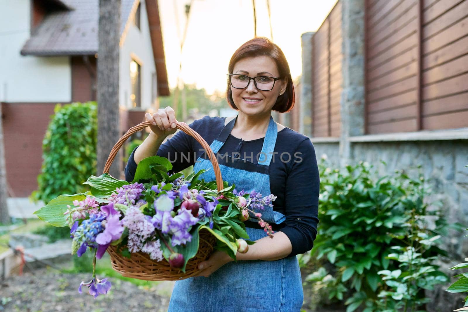 Middle-aged woman gardener florist in an apron with basket of fresh flowers folded in floral arrangement. Hobby, leisure, floristry, nature, beauty, gardening, creative bouquet, holiday concept