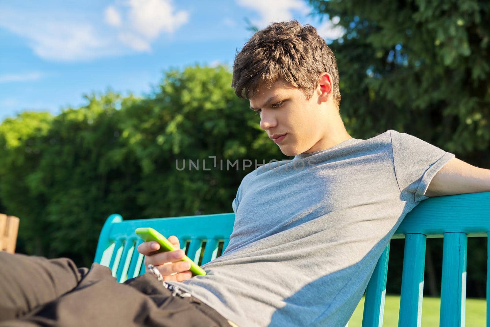 Serious guy teenager using smartphone, having rest, sitting on bench in park. Youth, technology, lifestyle, adolescence concept