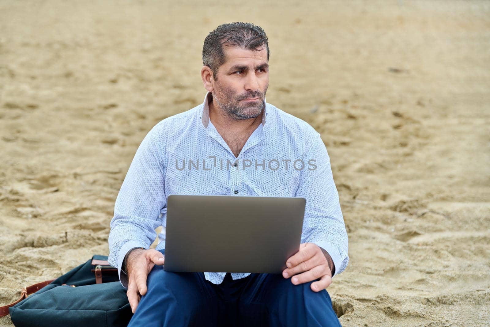 Serious confident mature man with laptop outdoors. Business male working with laptop sitting on sand at beach. Remote business, technology, freelance, lifestyle, leisure, middle age concept