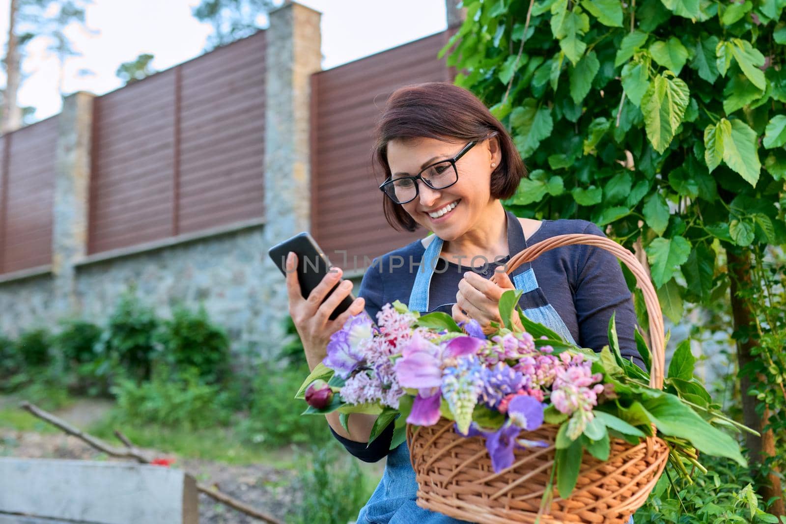 Middle-aged woman in garden with basket of fresh cut spring flowers using smartphone. Rest, gardening, lifestyle, leisure, emotions, spring, nature, technologies, middle age concept