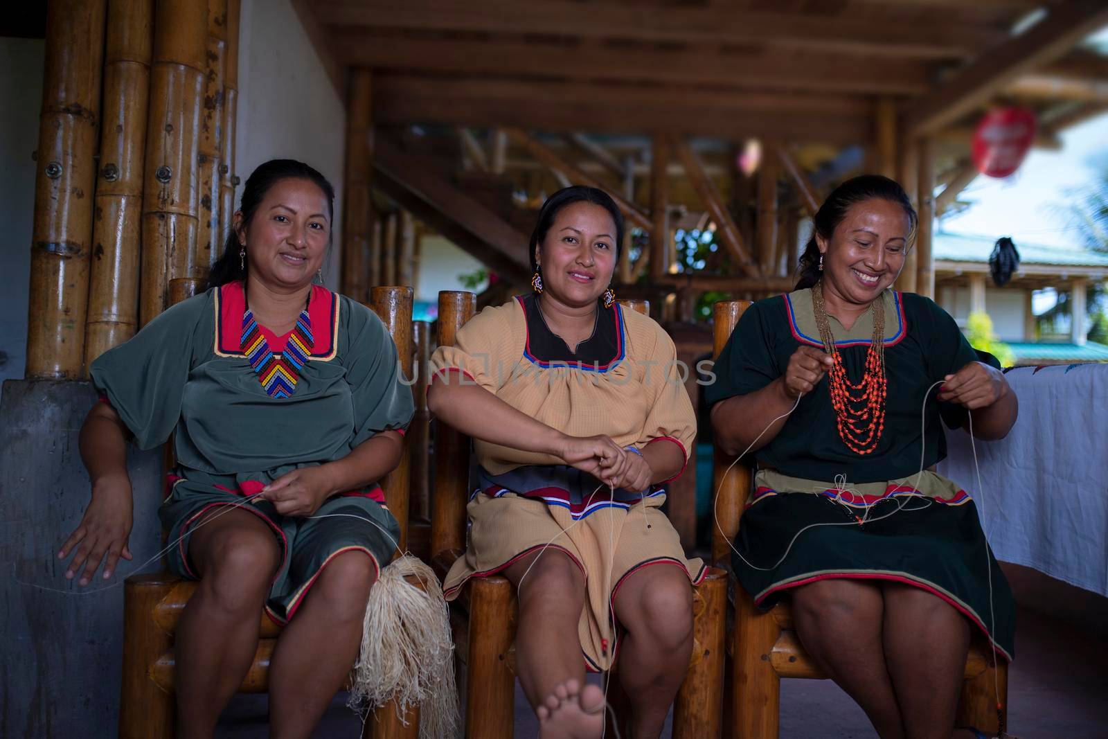 Indigenous woman of Cofan nationality with green dress smiling while weaving handicrafts sitting on a chair at her home in the Amazon rainforest by alejomiranda