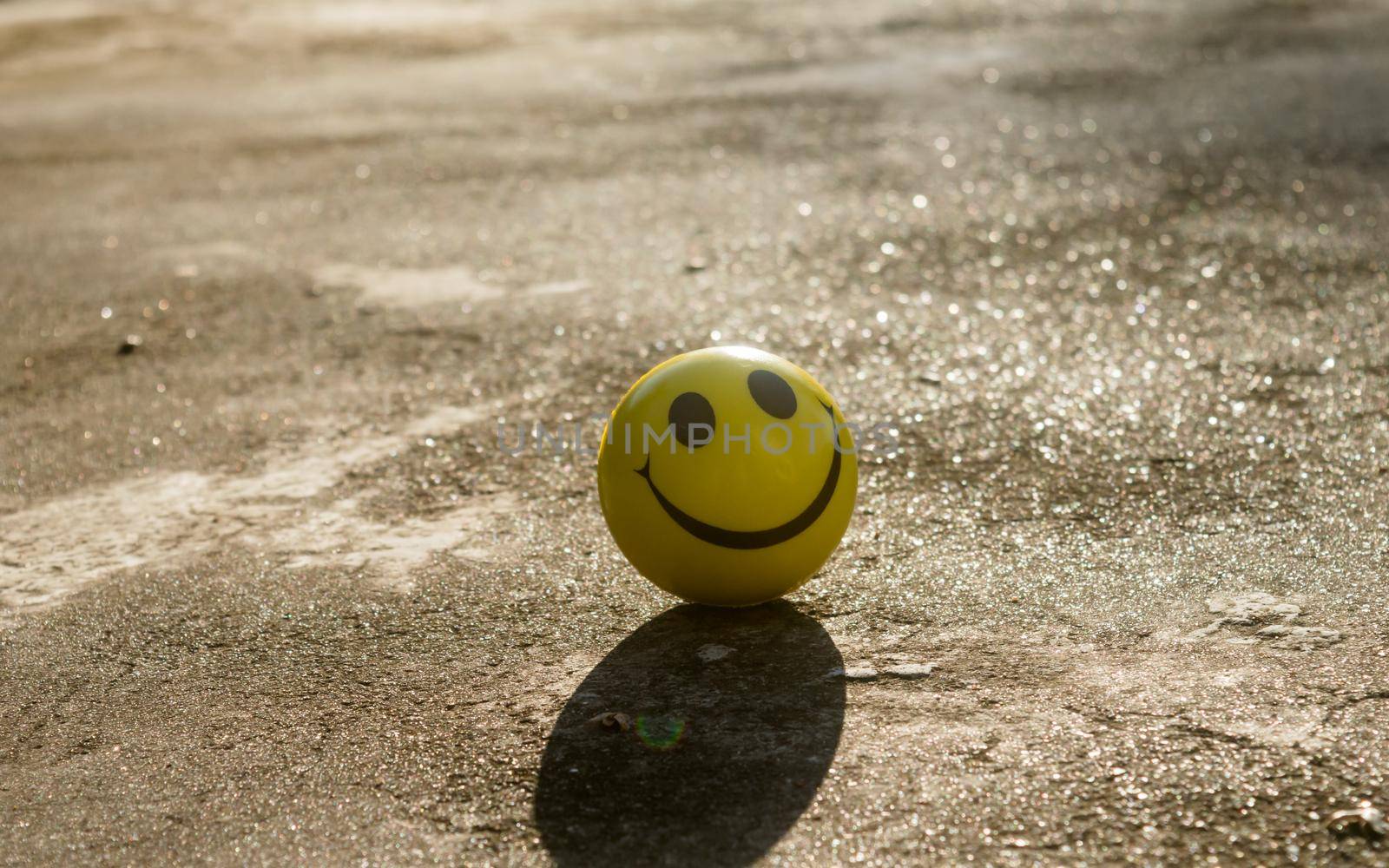 The iconic yellow smiley face with its perfect circle, two oval eyes and a large semi-circular mouth representing a symbol of happiness isolated on sand and shadow background. Happy smile backgrounds. by sudiptabhowmick