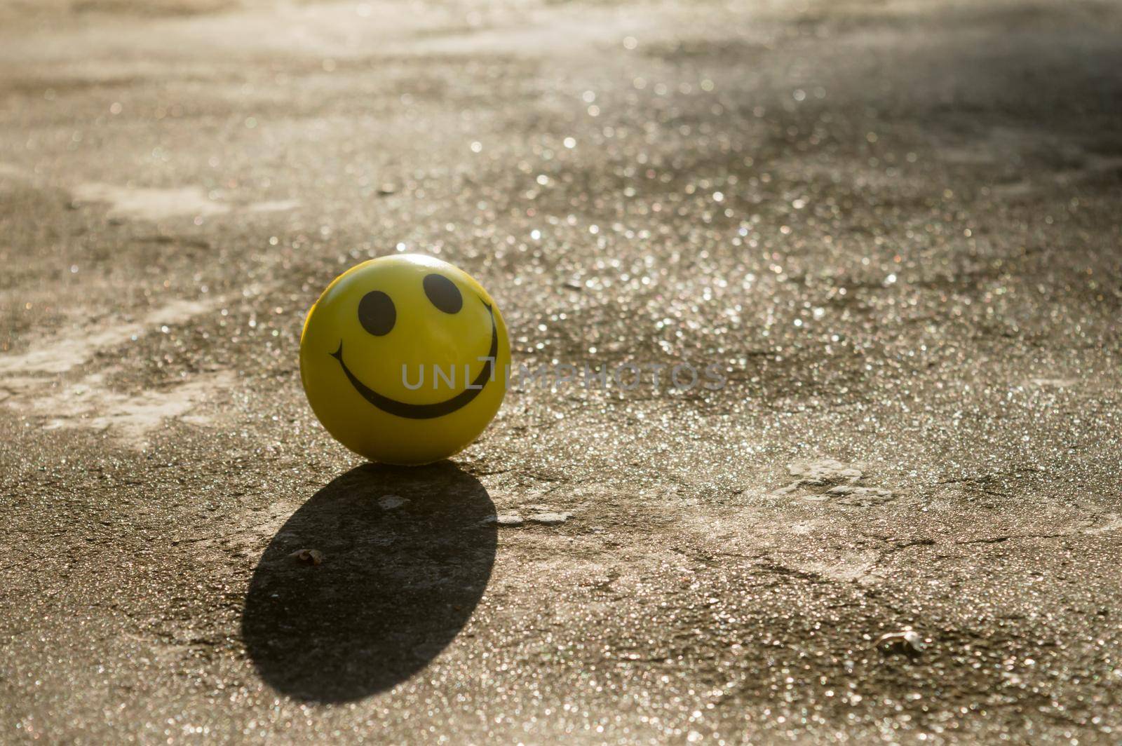The iconic yellow smiley face with its perfect circle, two oval eyes and a large semi-circular mouth representing a symbol of happiness isolated on sand and shadow background. Happy smile backgrounds. by sudiptabhowmick