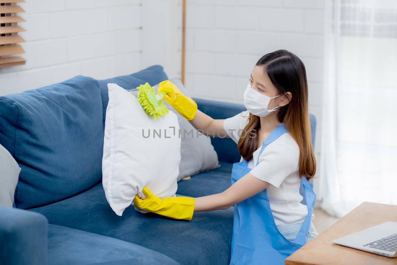 Young asian woman in face mask and gloves cleaning dust with duster on sofa and cushions at home, girl doing housework for hygiene at house, housekeeper and service, health care and lifestyle concept.