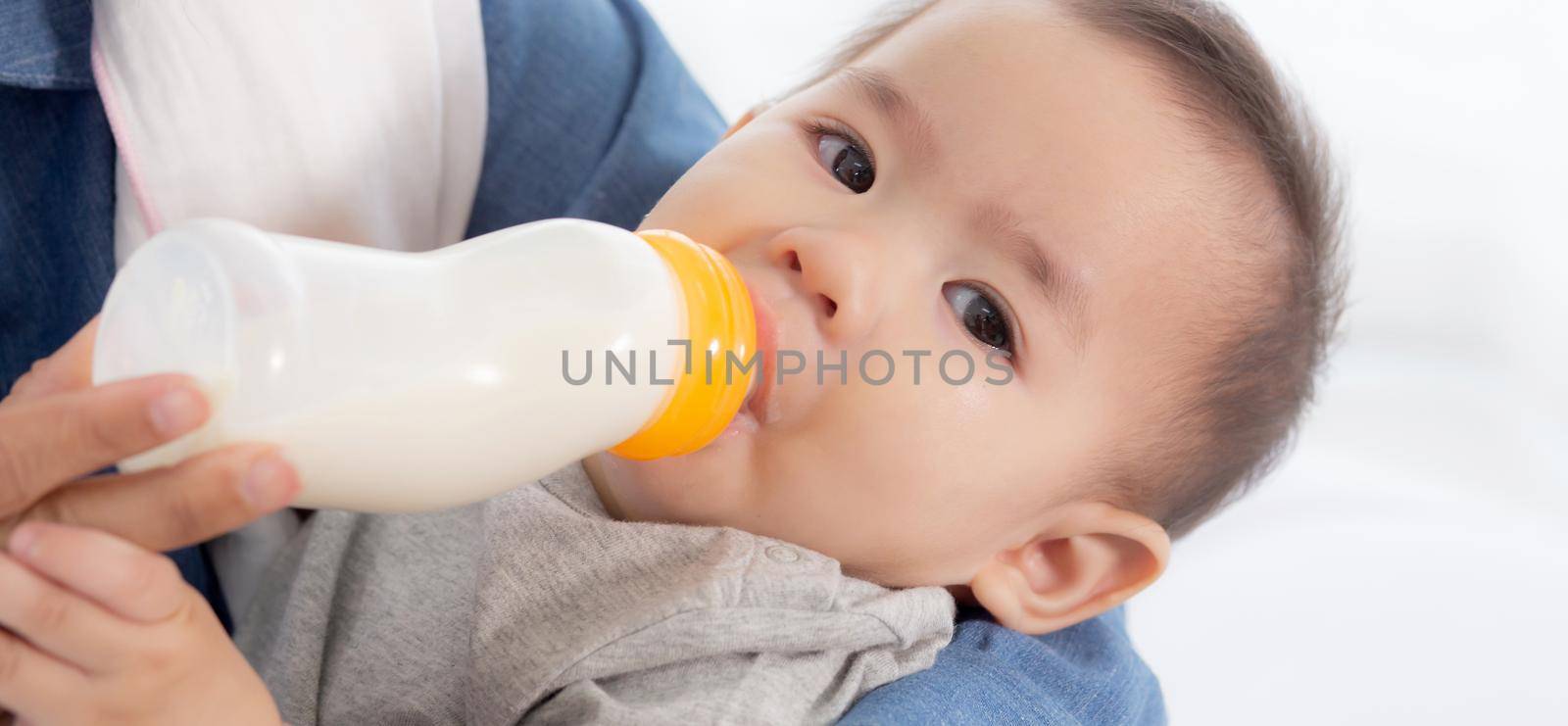 Young asian mother embracing and feeding little baby girl with bottle of milk at home, newborn innocence drinking with mom satisfied, relationship and bonding of mum and child, family concept.