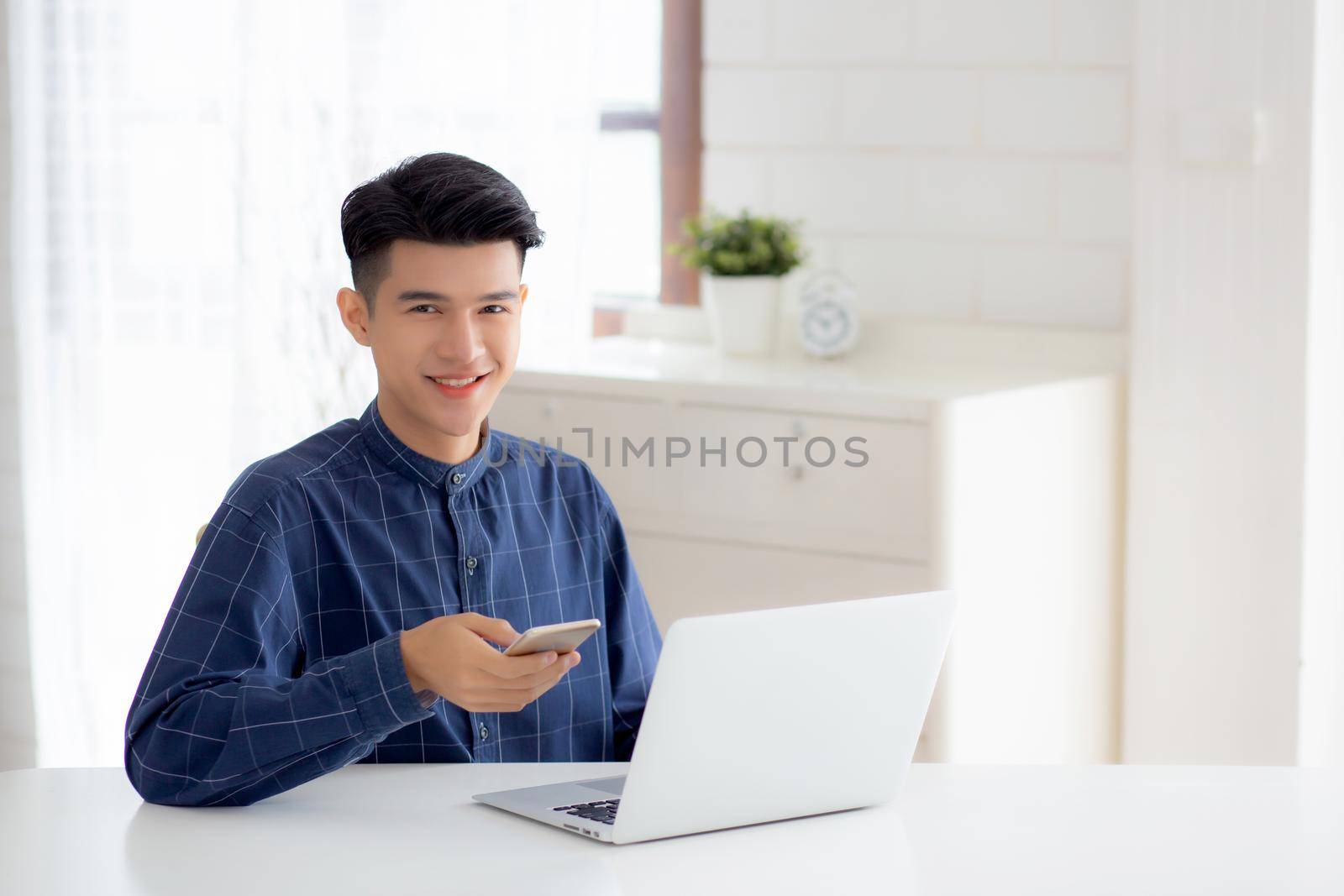 Young asian man working laptop computer and reading smartphone on internet online on desk at home, freelance male smiling using phone with social media on table, business and communication concept.