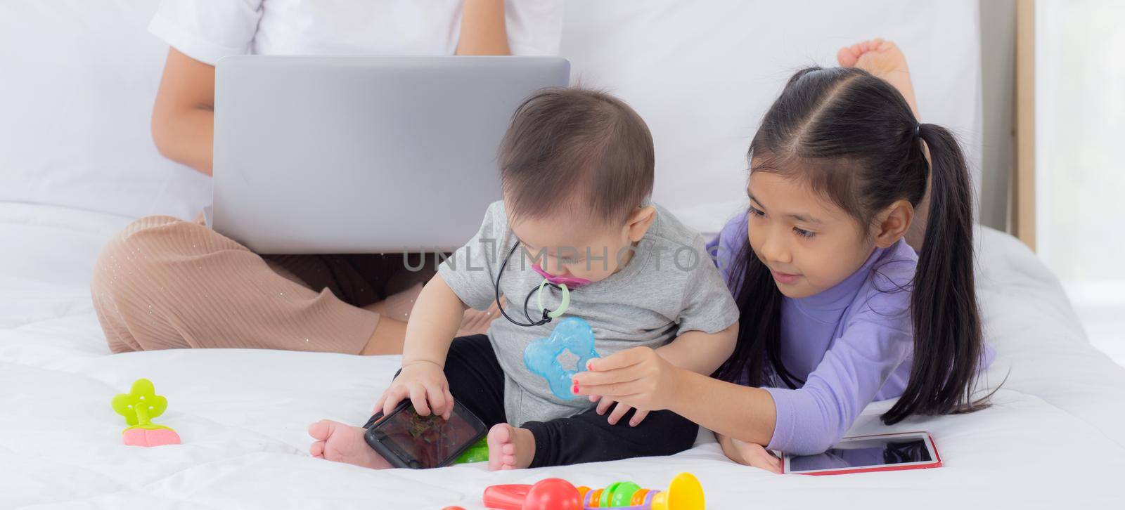 Young asian mother working at home online to internet with laptop computer and talking phone and daughter looking tablet on bed at bedroom, business woman is freelance, new normal, family concept.