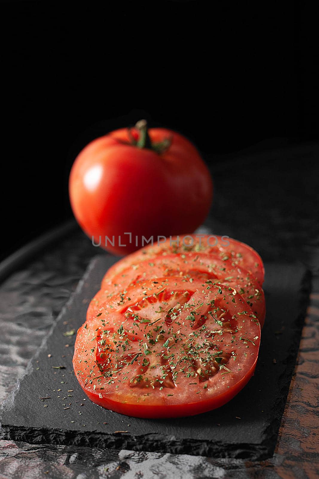 Sliced ​​and whole tomatoes with herbs. Black cutting board.
