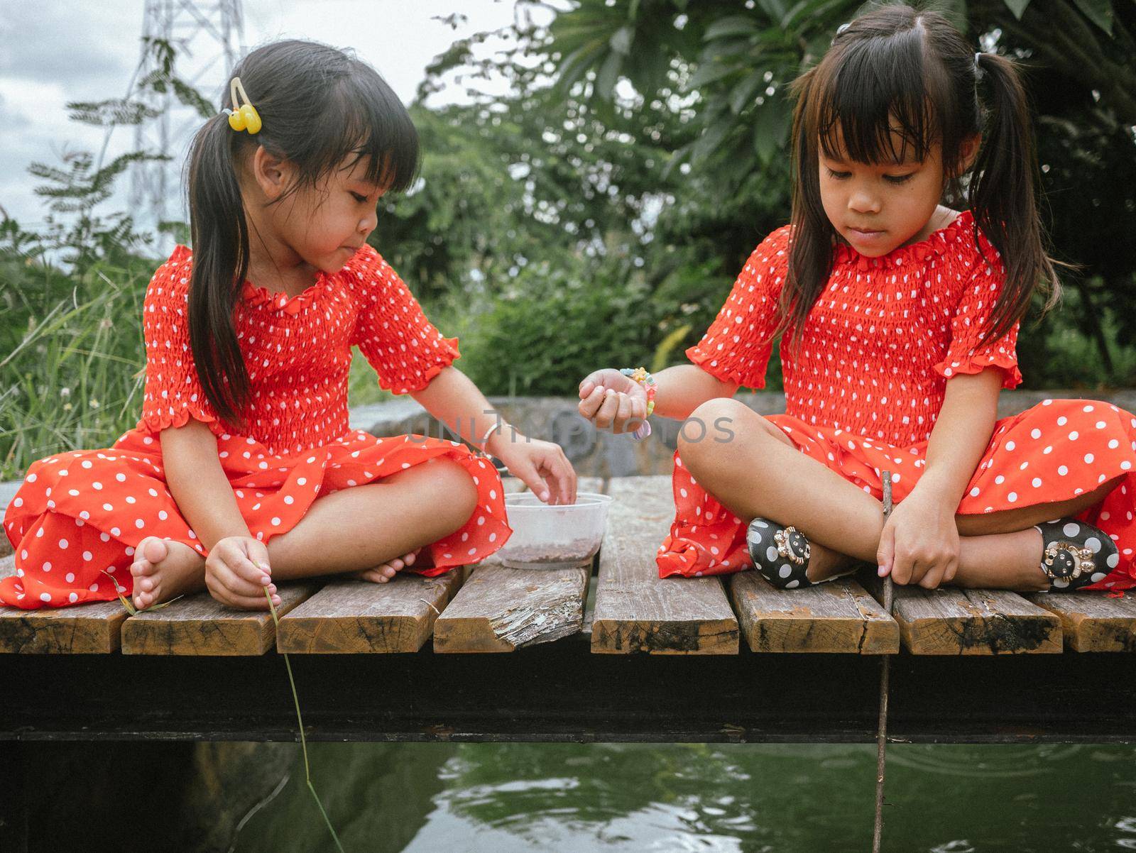 Two little sibling girls in red dress role playing fishing sitting on a wooden bridge by the pond. Childhood happiness. by TEERASAK
