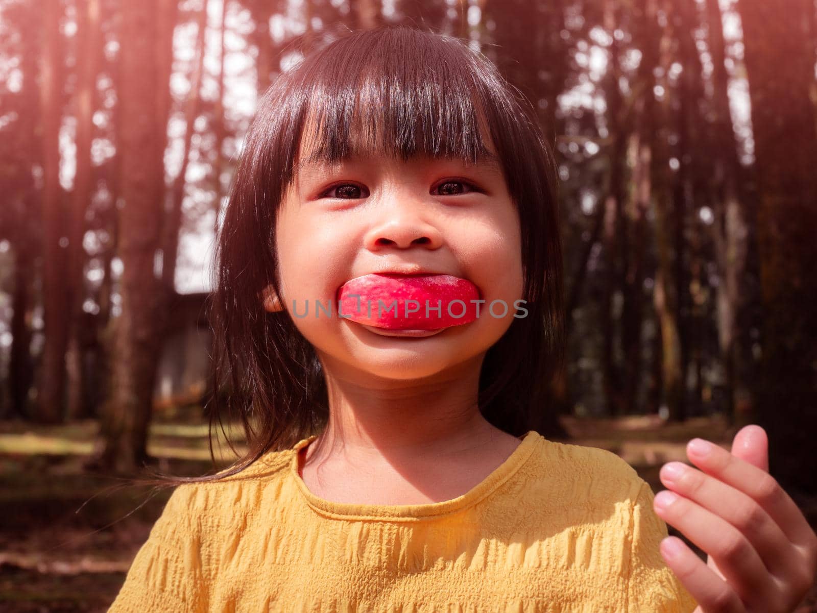 Portrait of happy little girl eating red apple outdoors in summer. Childhood happiness. by TEERASAK