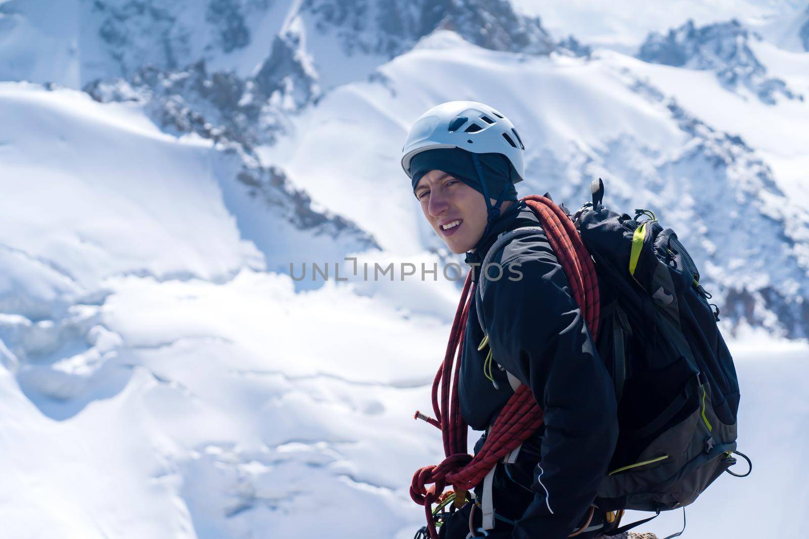 A young man traveler is engaged in mountaineering. Hiker in a helmet, with a rope climbs to the top, against the backdrop of a stunning view with snow-capped mountains.