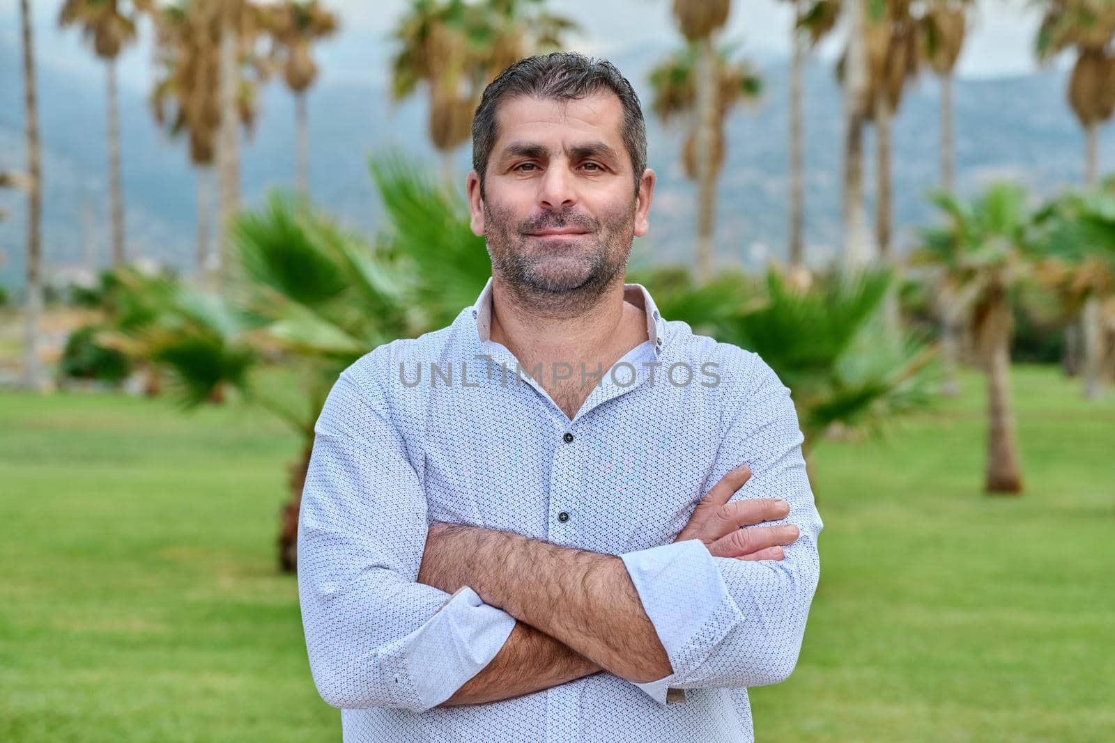Portrait of confident serious mature man outdoor. Middle aged male looking at camera with crossed arms, tropical park background. Age, lifestyle, people concept