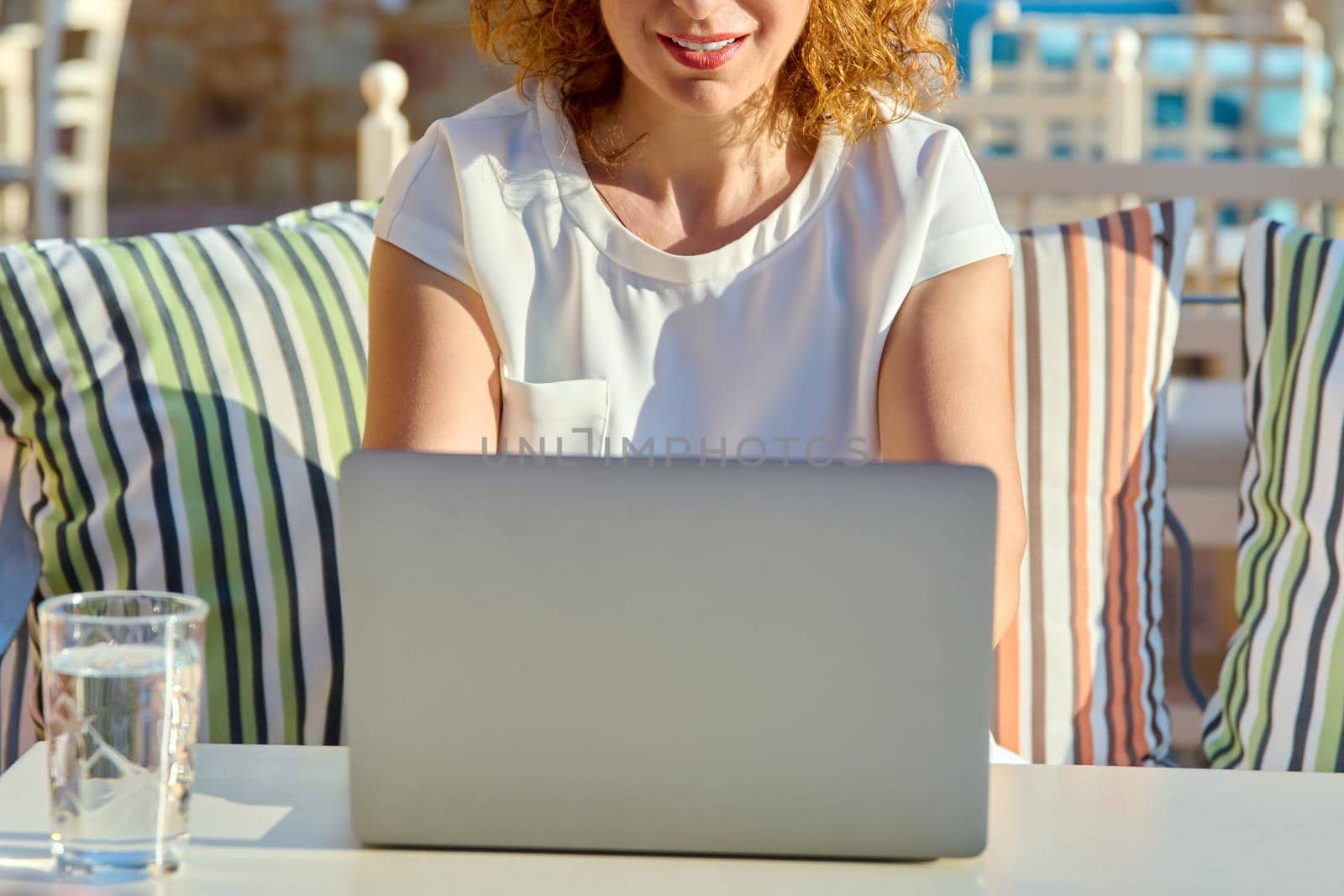 Positive smiling woman working with laptop outdoors. Middle aged female with headphones sitting at table in cafe, looking at monitor. Remote work, freelance, online business, video conference, chat