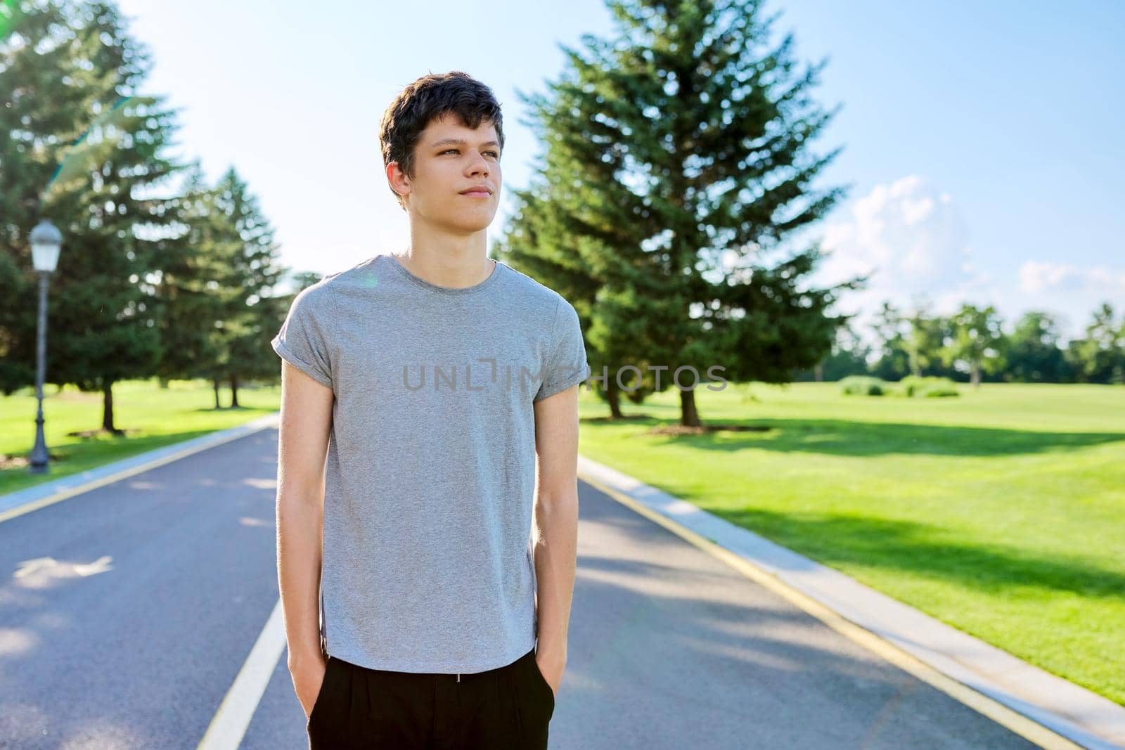 Portrait of a smiling male teenager looking at the camera, guy on the road in a summer green park. Adolescence, lifestyle, recreation and leisure youth concept