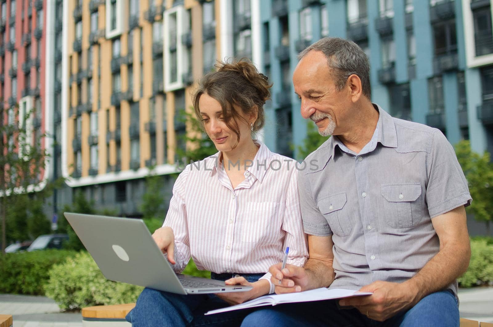 Attractive young woman and senior man are using laptop sitting on the bench in modern residential complex. Casual meeting outside office. A daughter is teaching technology for her father by balinska_lv